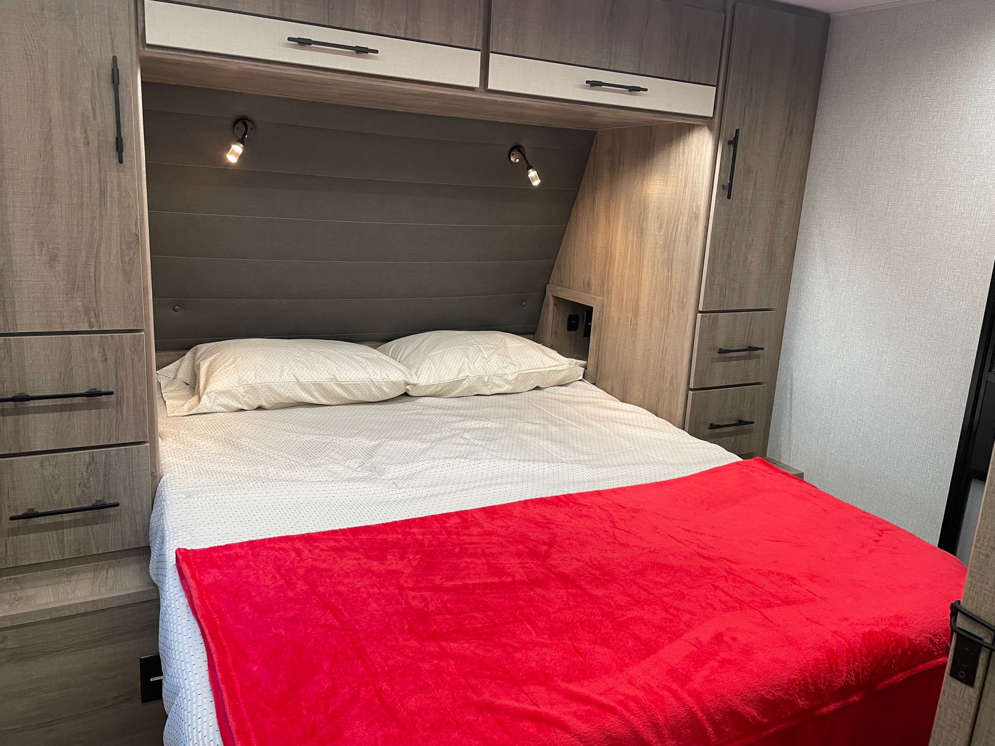 Comfortable RV rental bedroom featuring built-in storage, reading lights, and a cozy red blanket for travel.. Grand Design Imagine XLS 2024