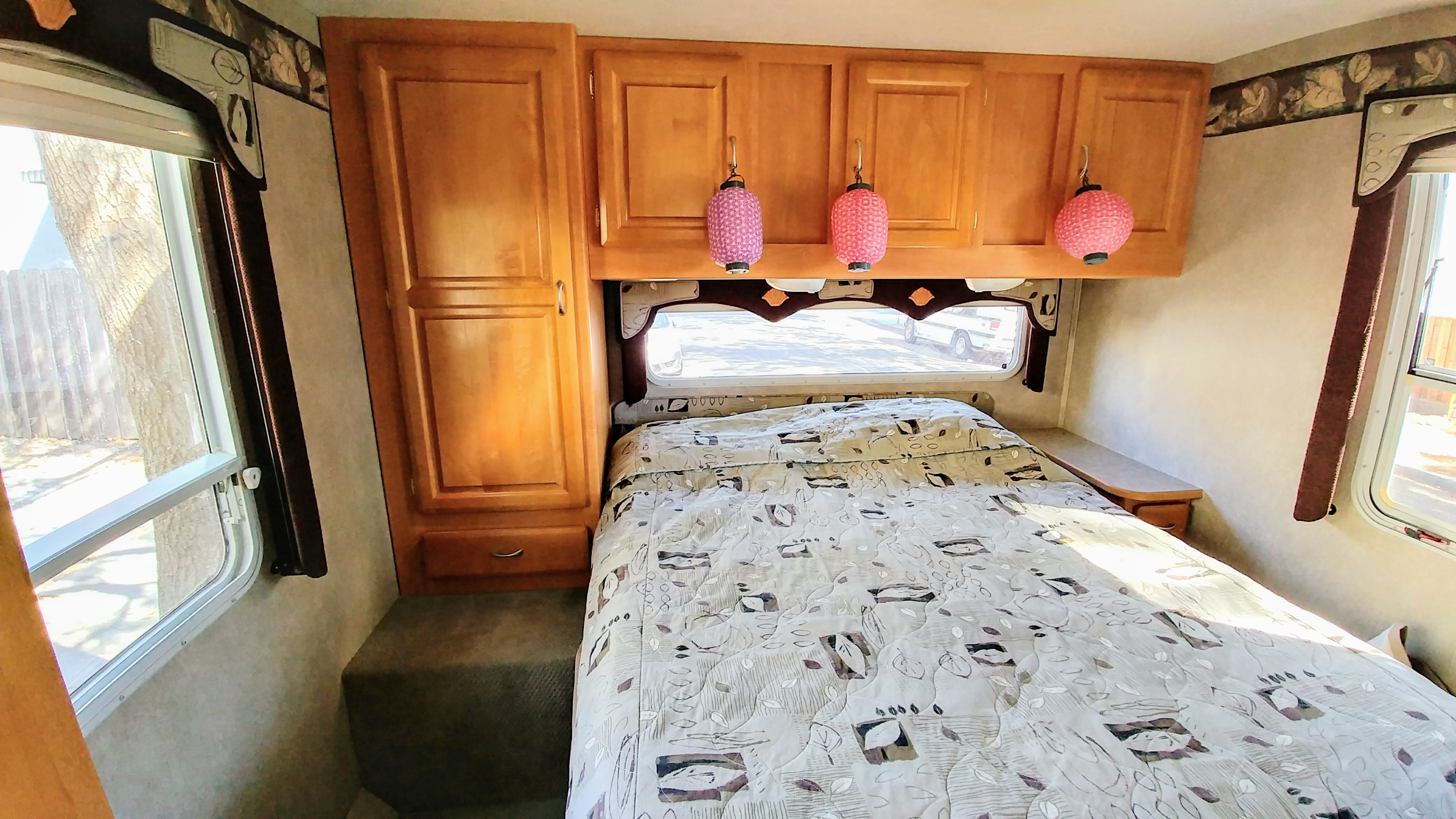 Comfortable RV rental bedroom with a patterned bed, wood cabinets, and three pink lanterns. Bright windows illuminate the space.. Jayco Greyhawk 2005