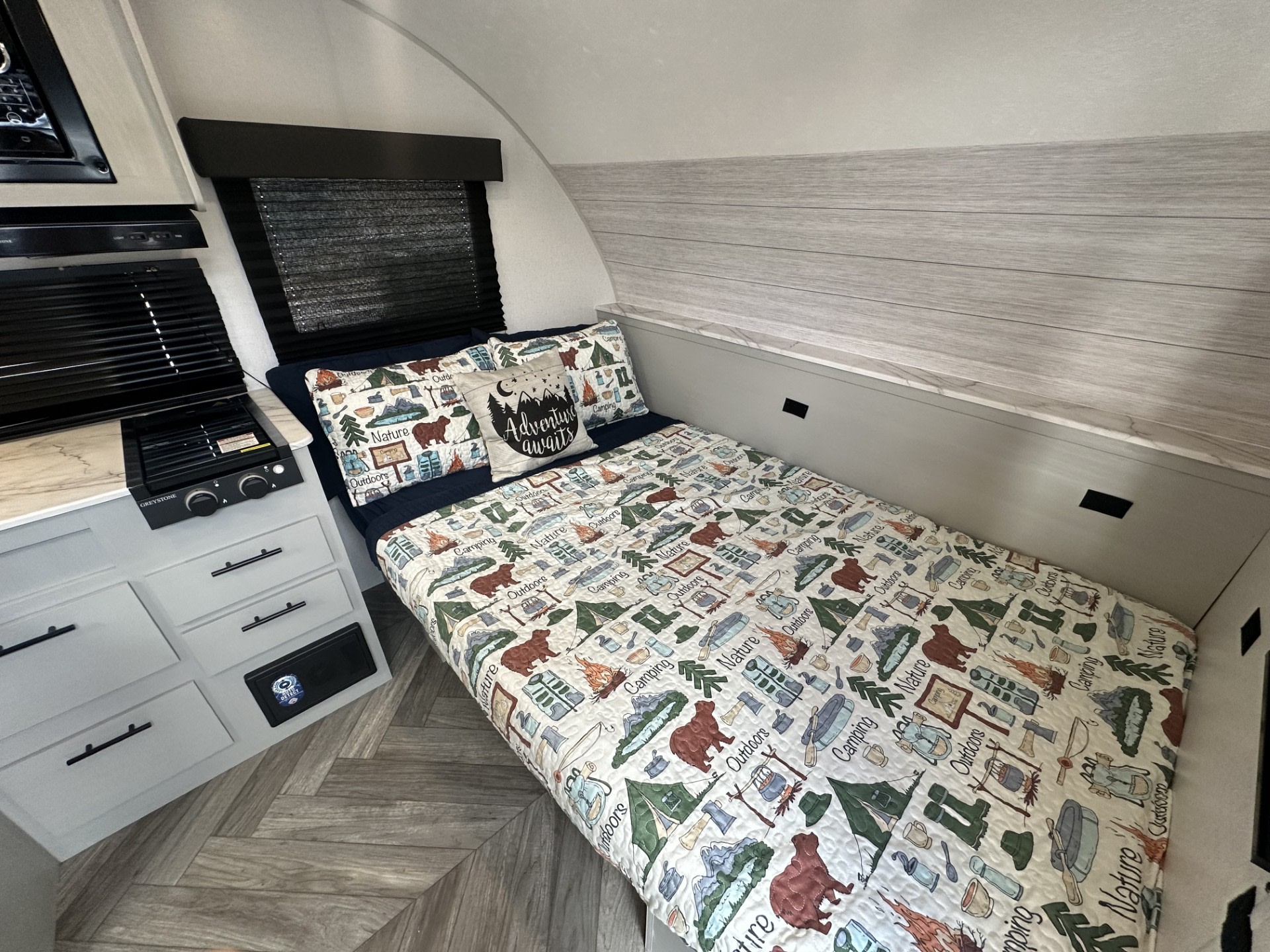 Inside a compact rv rental: a cozy bed with camping quilt & "Adventure Awaits" pillow, next to a small kitchenette.. Forest River Wildwood 2022