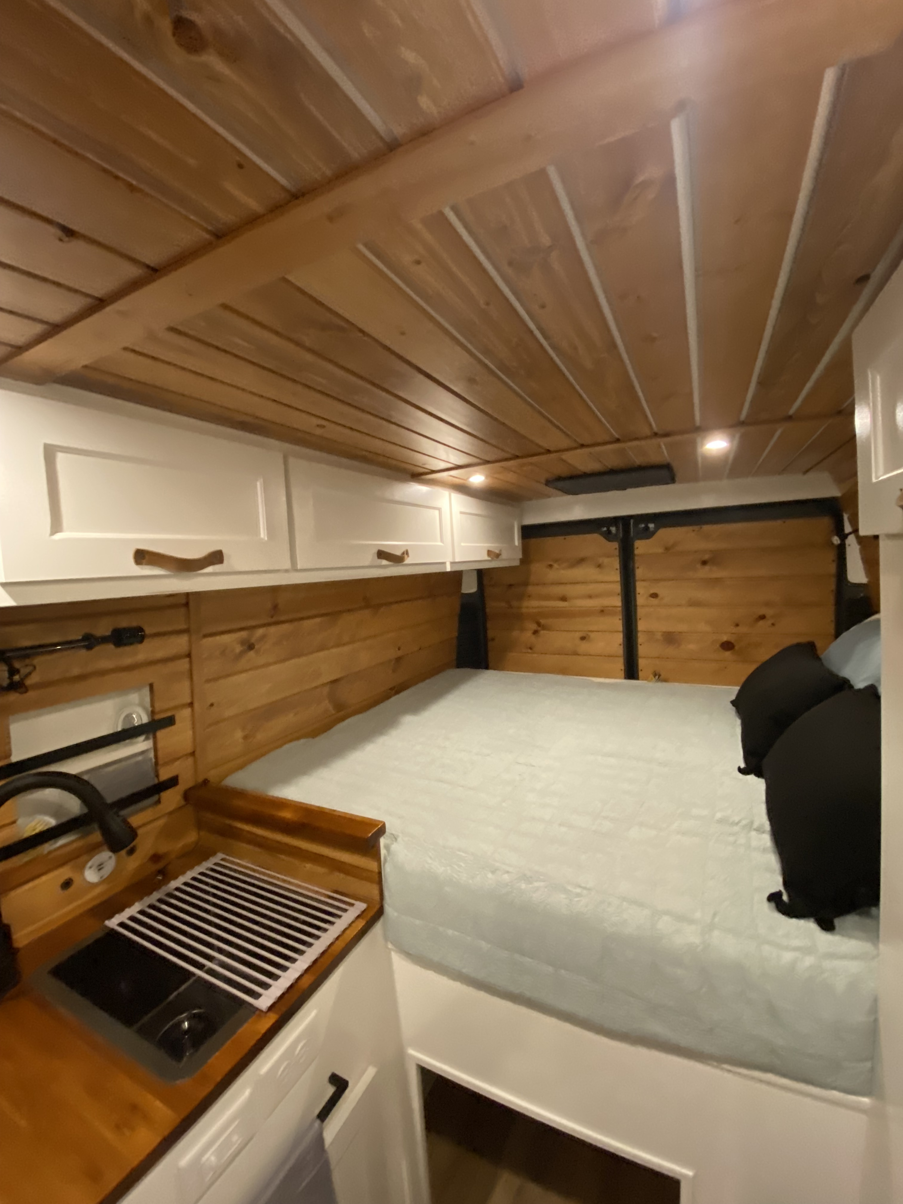 Well-appointed RV rental camper van interior. Shows a comfortable bed, wood-paneled walls/ceiling, white cabinets, and a kitchen sink.. RAM Promaster 2300 2023