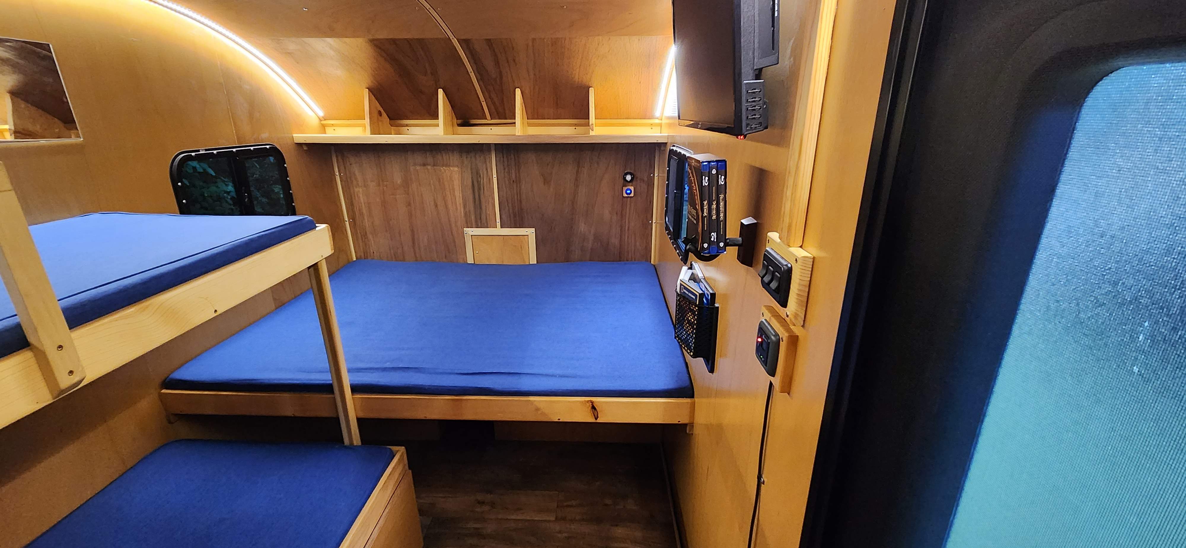 This RV rental's compact wooden interior features bunk beds, a main bed with blue cushions, LED lighting, and a TV setup.. Bend Teardrop Tall Boy 2023