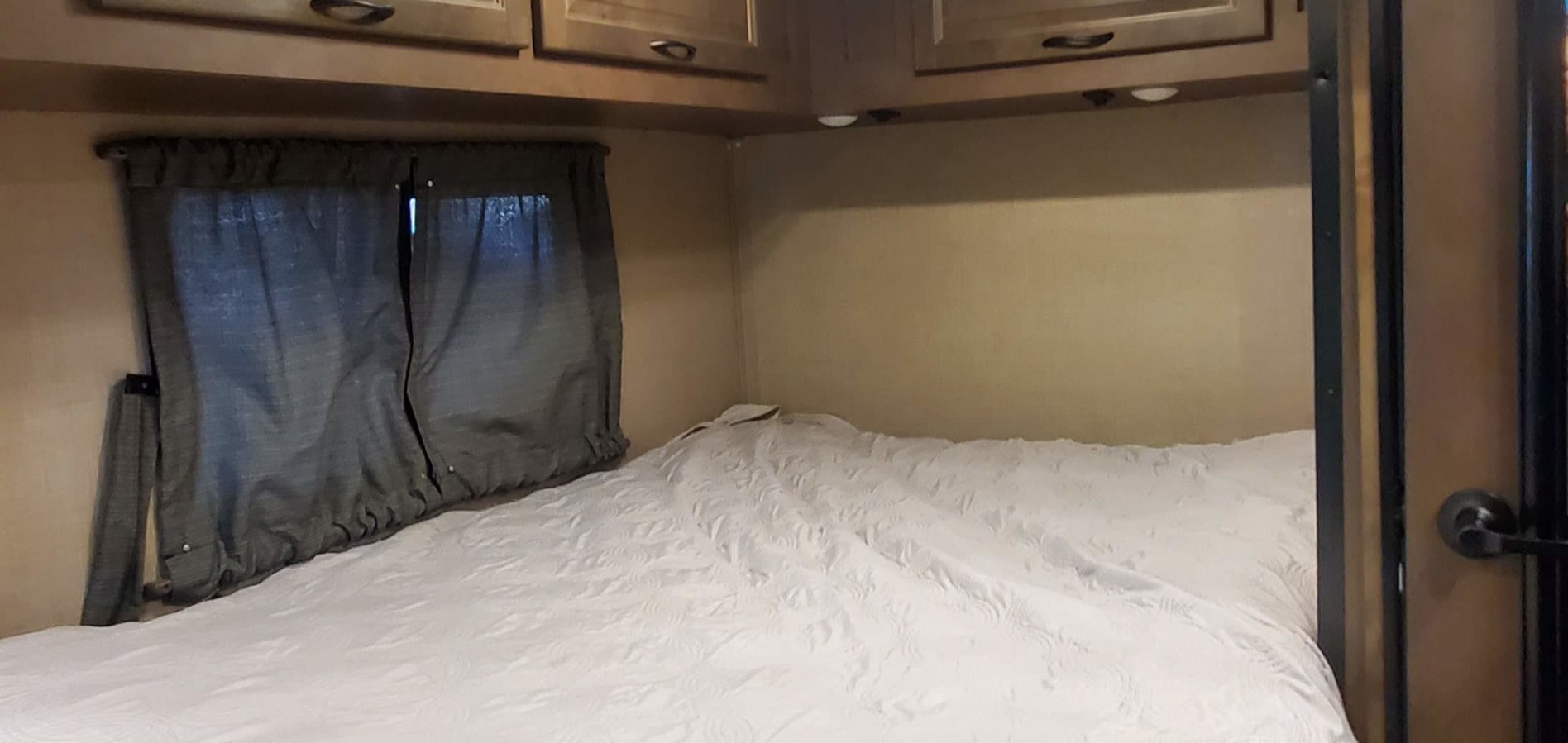 Cozy RV rental bedroom showing a white bed, closed grey curtains, and wooden overhead cabinets for comfortable travel.. Wayfarer T23 2020
