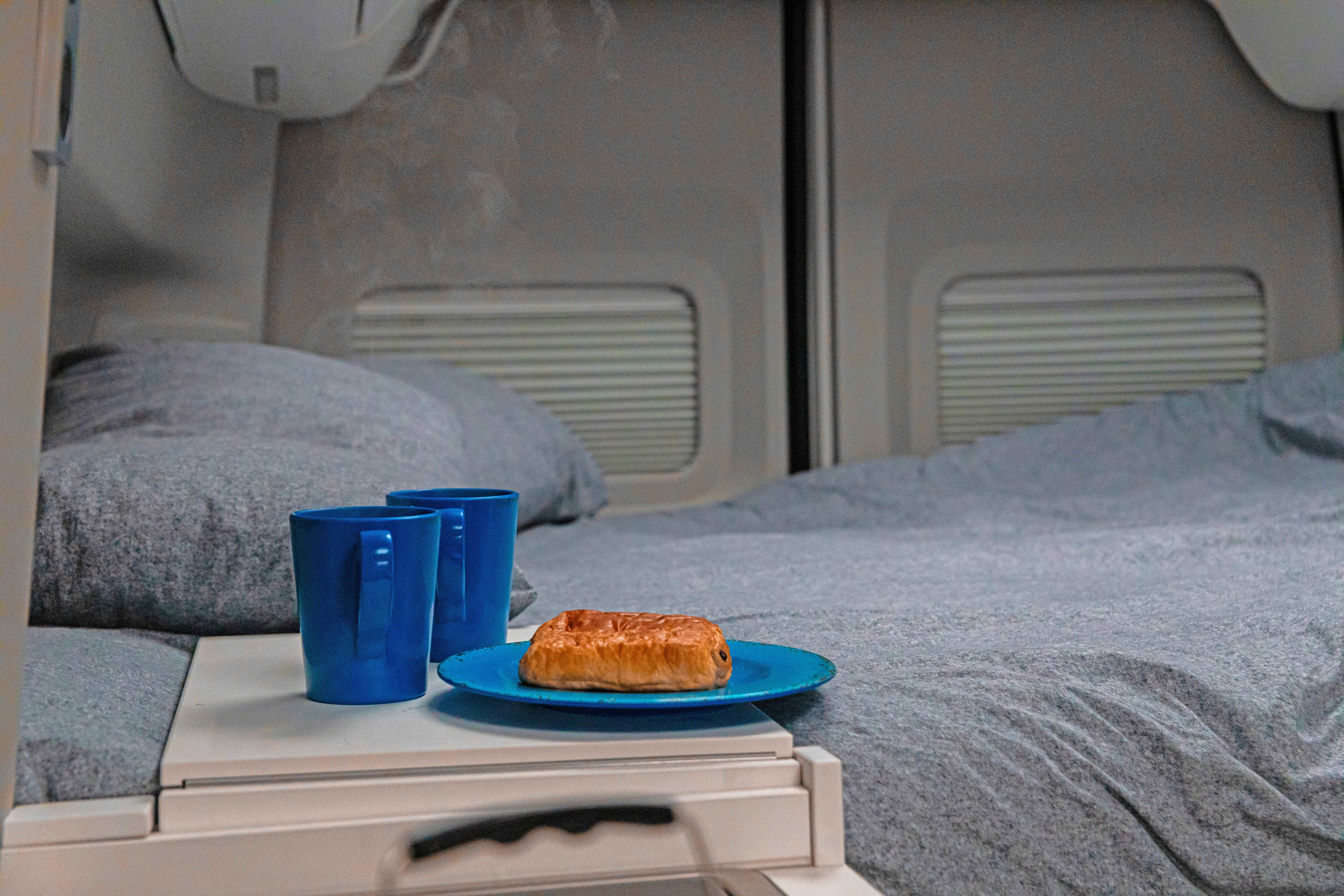 Cozy RV rental breakfast: steaming drinks in blue mugs, pastry on plate by a gray bed.. Volkswagen Grand California 2024