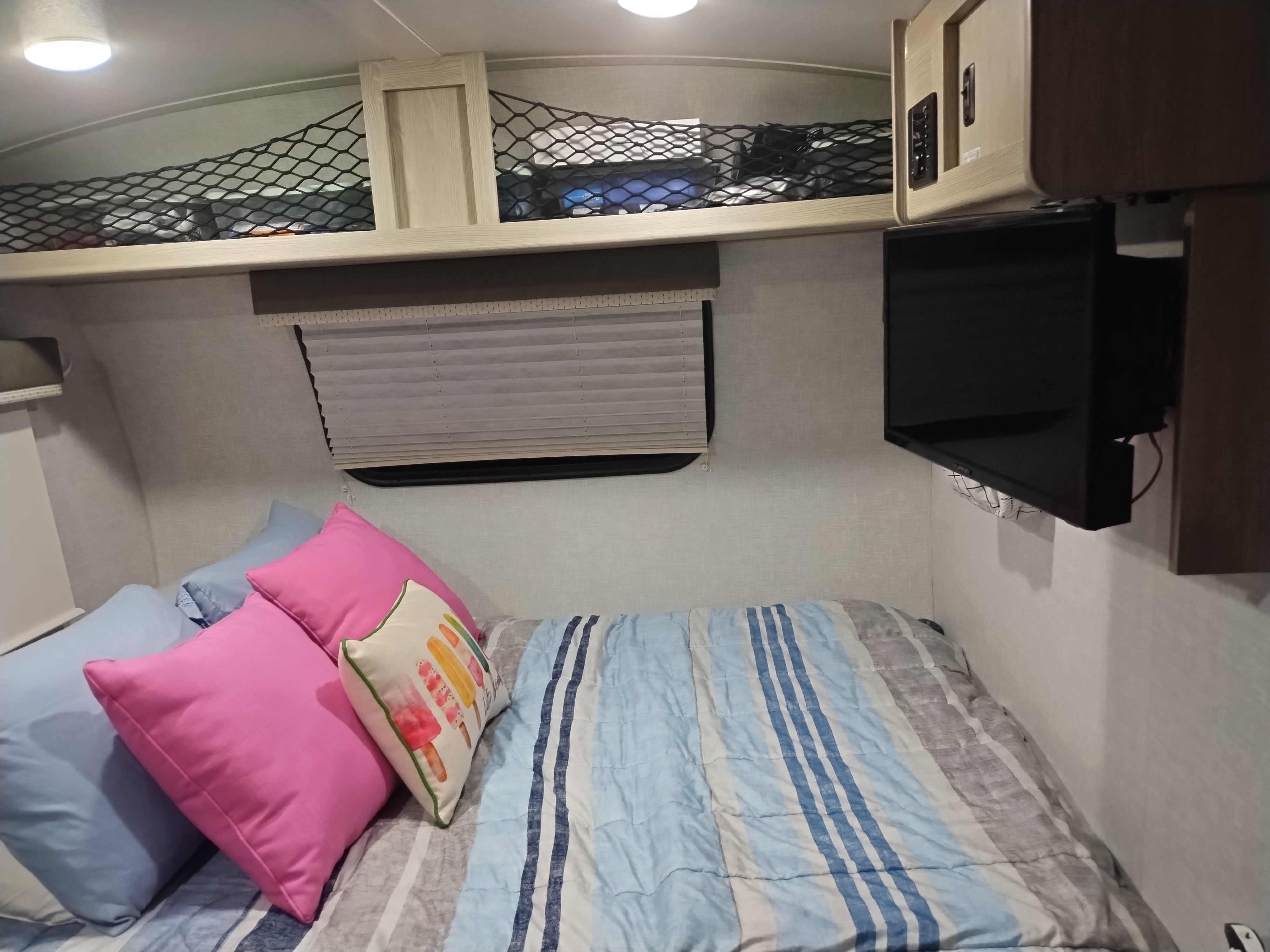 Cozy RV rental bedroom featuring a bed with colorful pillows, overhead storage nets, a mounted TV, and window blinds.. Forest River Rockwood Geo Pro 2021