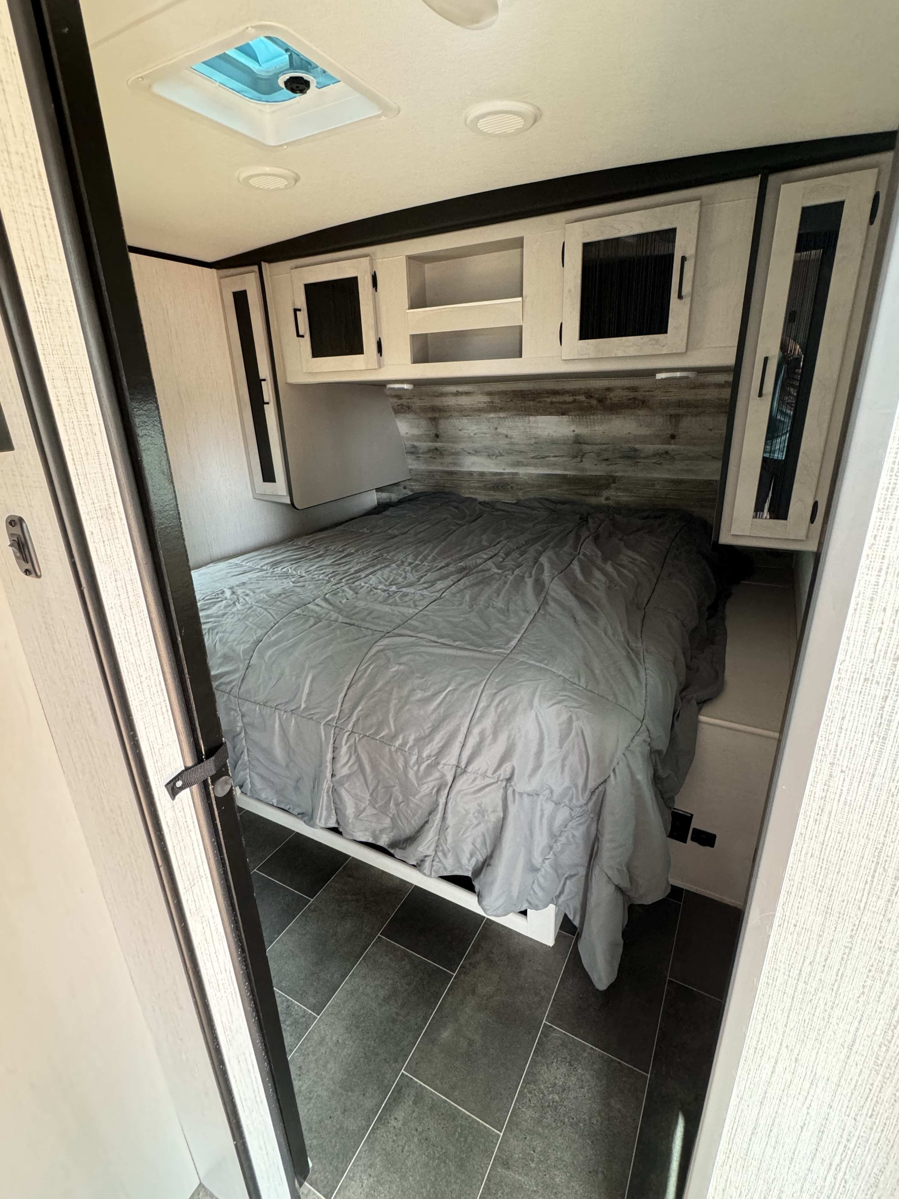 Clean RV rental bedroom: gray bed, overhead cabinets, wood-look accent wall, and dark tiled floor.. Heartland RVs Mallard 2022