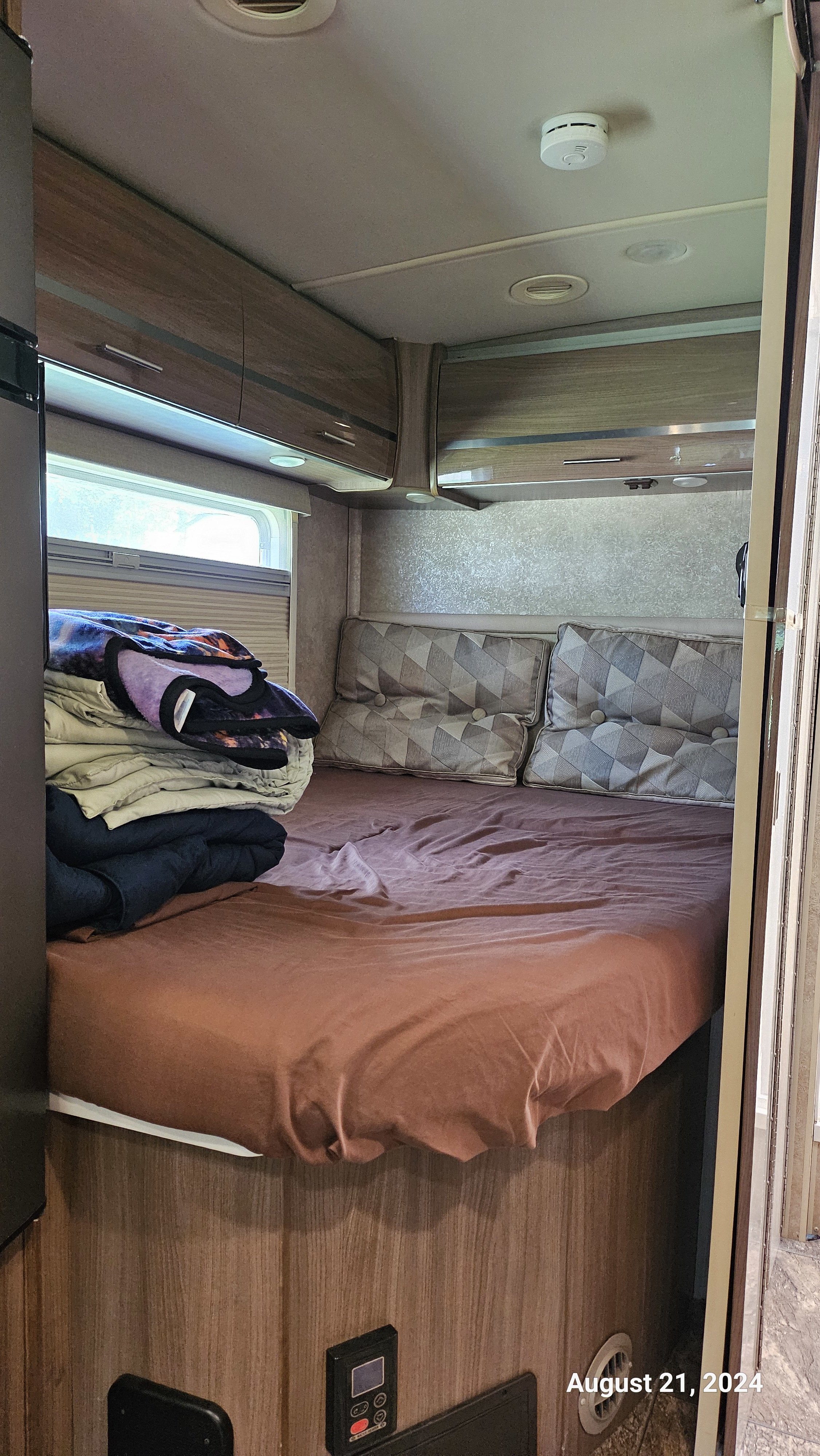 Interior view of a compact RV rental bedroom, featuring a bed, overhead cabinets, and a window for travel comfort.. Winnebago Via 2014