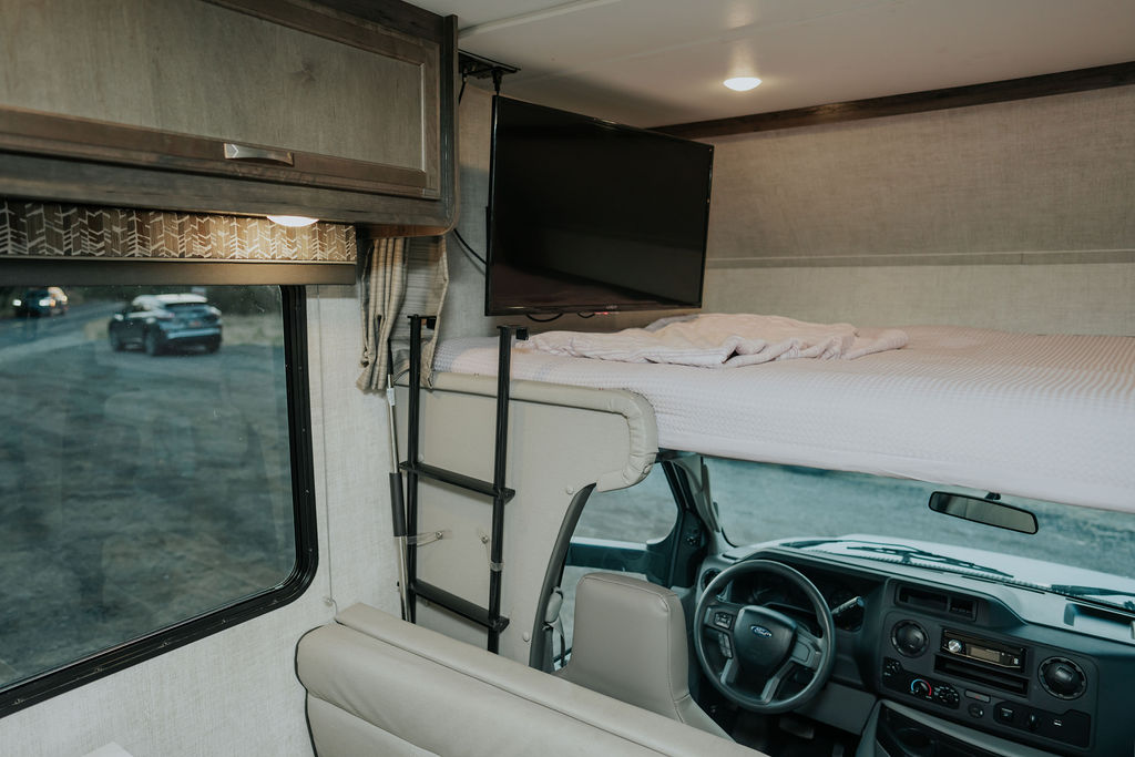 Inside an RV rental, showing the cab-over bed with a TV, ladder, and driver's area. Your spacious home on wheels.. Gulf Stream Conquest LE 2023