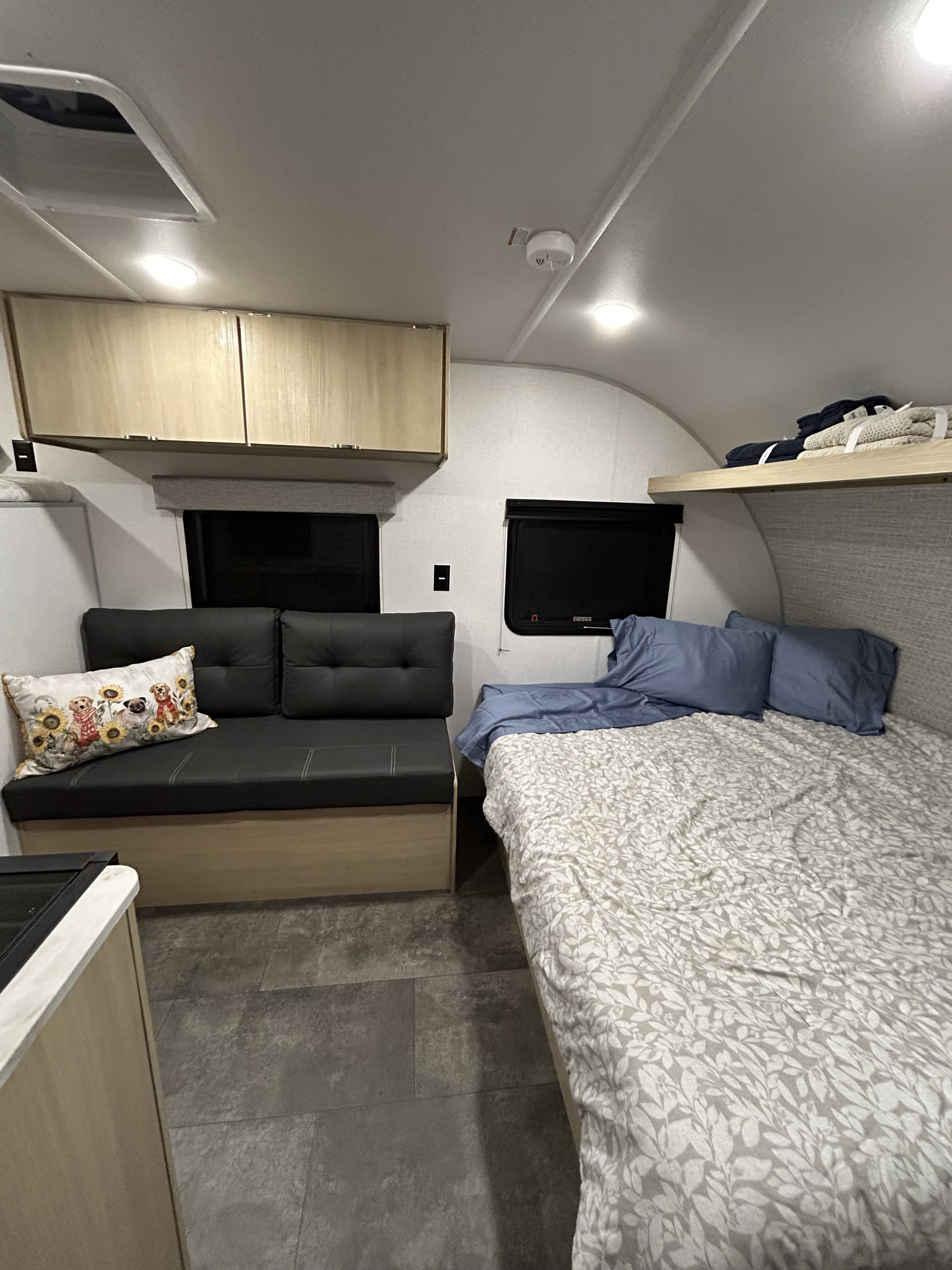 Inside an rv rental: a comfy bed, dark sofa with a fun dog pillow, light wood cabinets, and gray floor.. Wildwood 174BHLE 2025