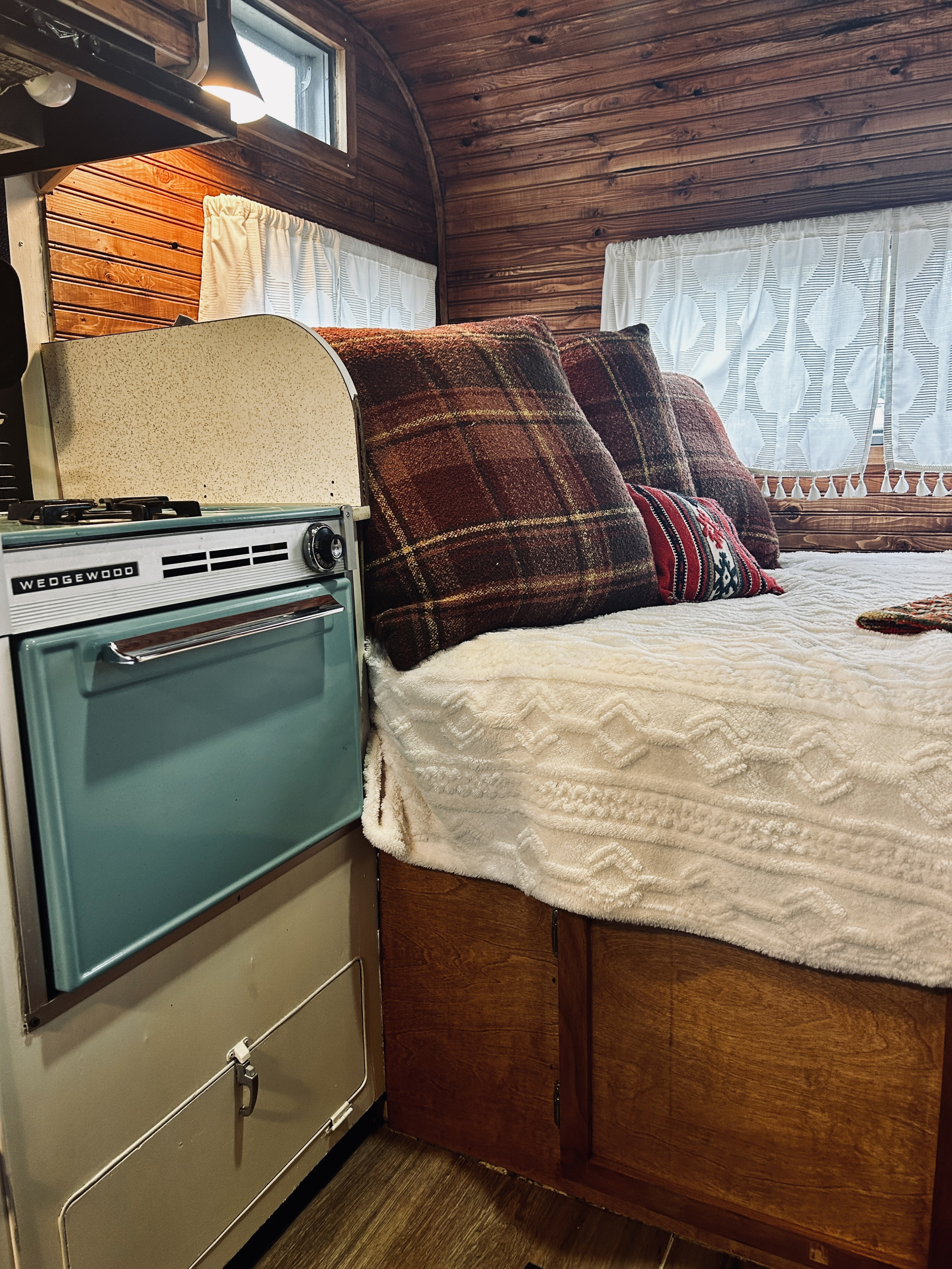 Cozy RV rental interior featuring a vintage Wedgewood stove, plush bed with plaid pillows, and warm wood paneling.. Skyline Nomad 1966