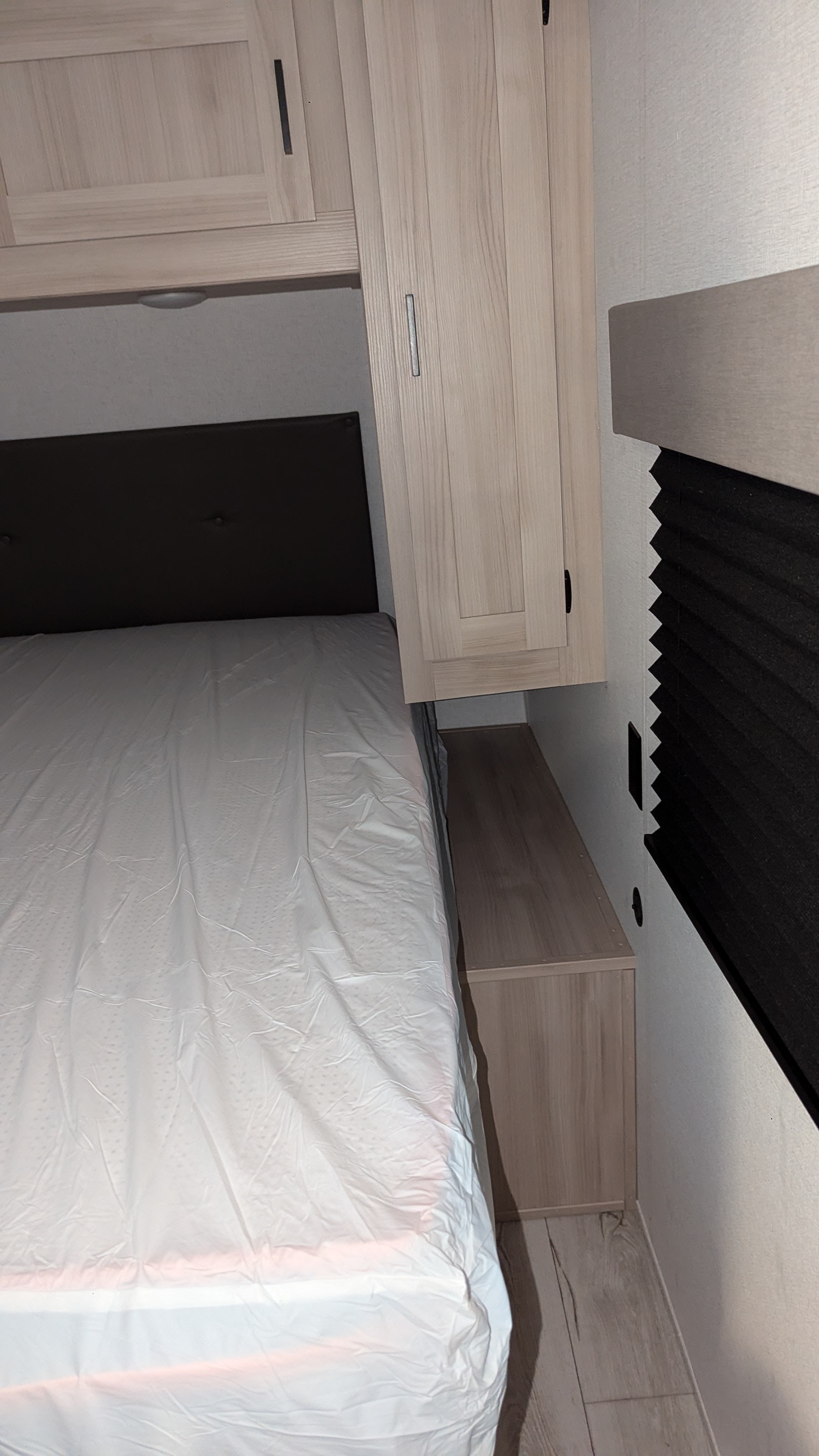 An RV rental bedroom features a bed with white protective cover, light wood cabinets, and window with pleated blinds.. East to West Della Terra LE 2024