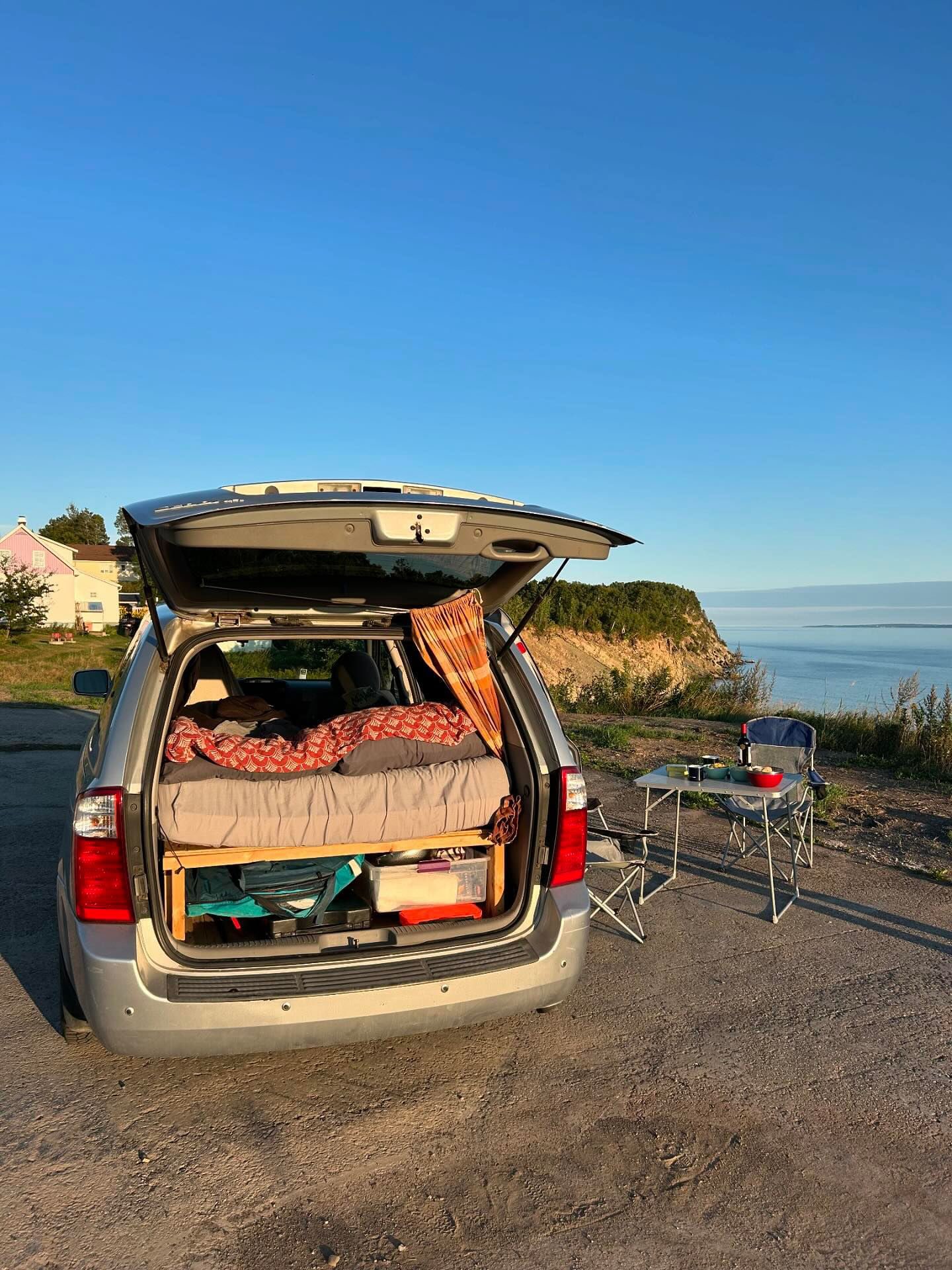 This silver minivan, like an RV rental, is converted for camping, featuring a bed and outdoor dining setup by a scenic ocean cliff.. KIA SEDONNA 2008