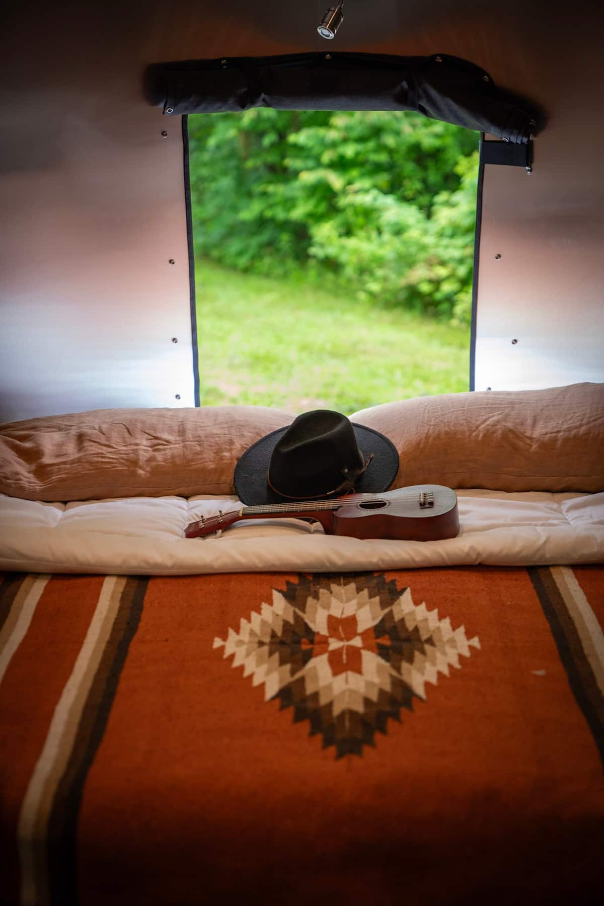 Relax in an RV rental: a cozy bed with a hat and ukulele, offering a view of lush green outdoors. Your perfect mobile home getaway.. Airstream Base Camp 2023