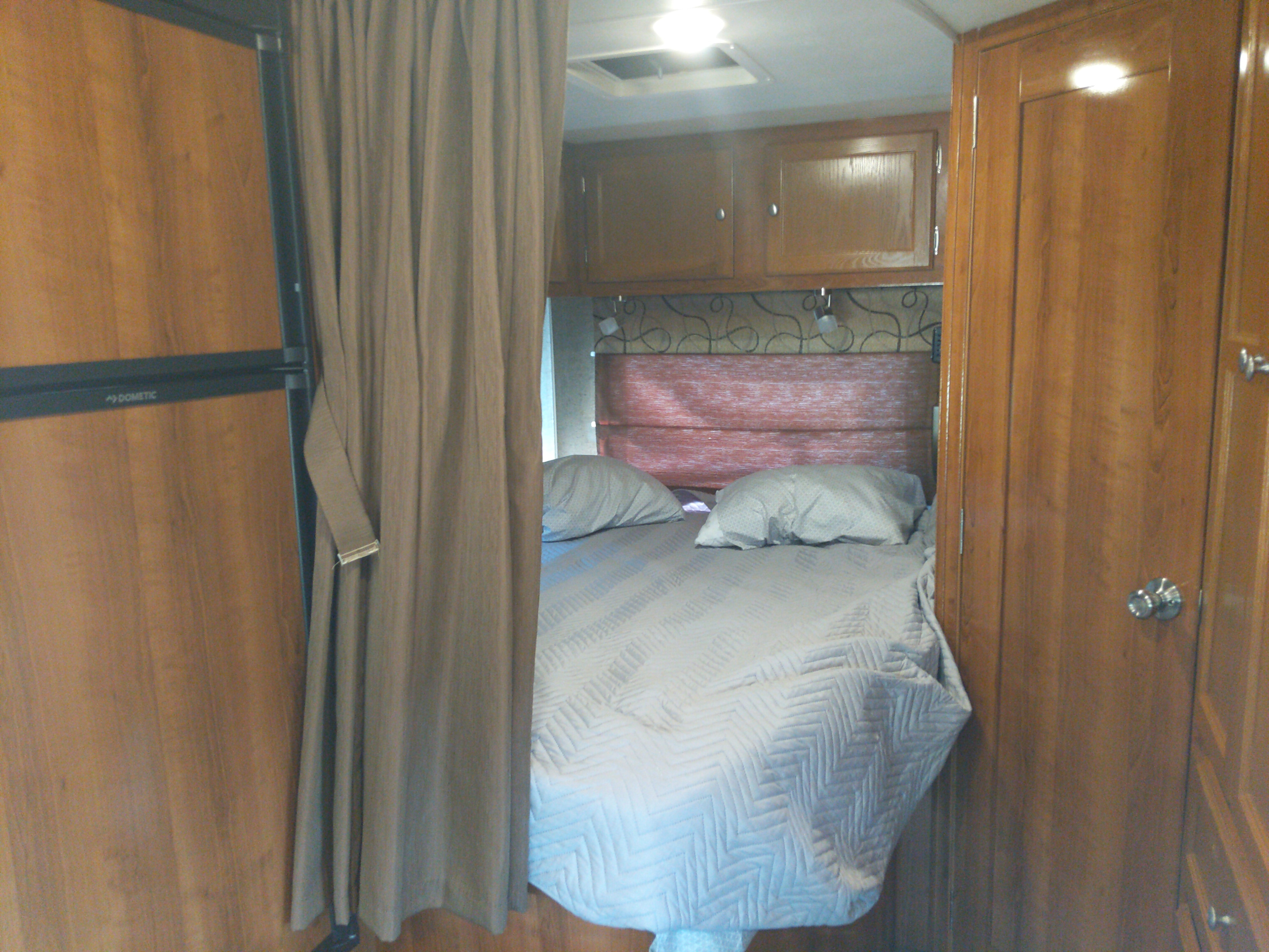 RV rental interior: A cozy bedroom space with a bed, pillows, and privacy curtain, plus an adjacent Dometic fridge.. Coachmen Leprechaun 230 CB 2018