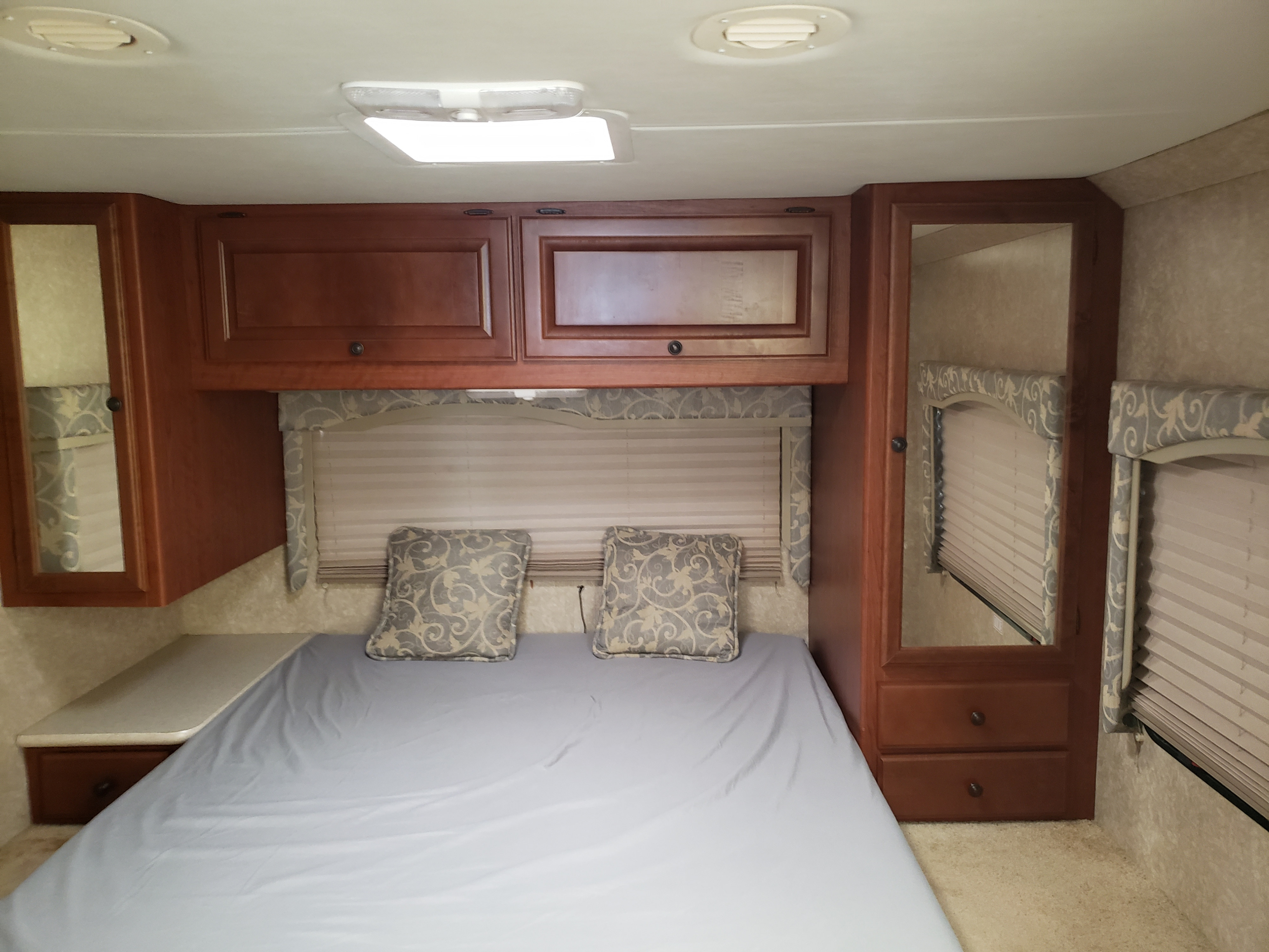 Detailed RV rental bedroom featuring a queen bed, rich wooden cabinetry, reflective mirrors, and shaded windows.. Four Winds Freedom Elite 2010