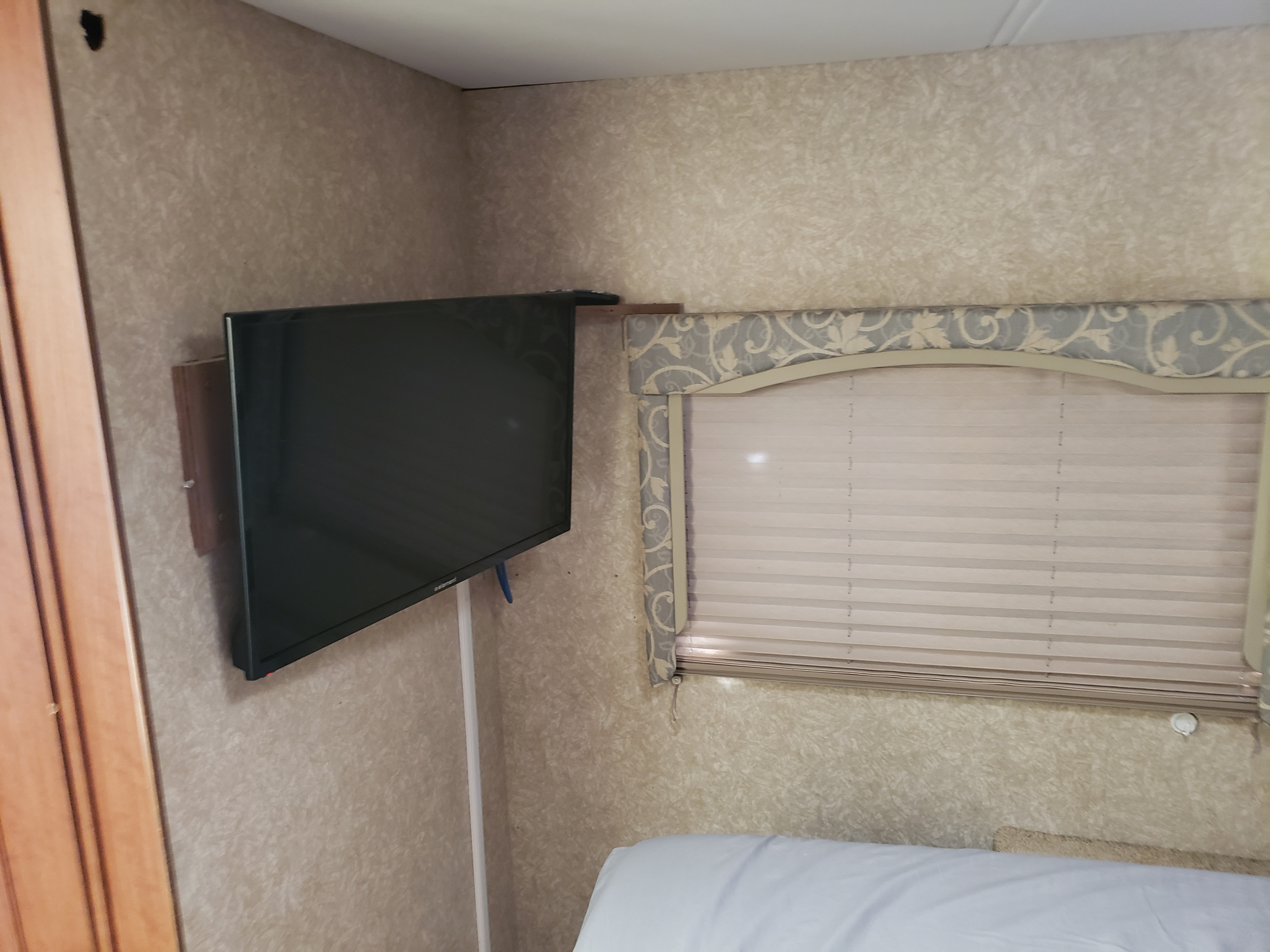 RV rental interior with a wall-mounted TV, decorative window valance over blinds, and a glimpse of the bed.. Four Winds Freedom Elite 2010