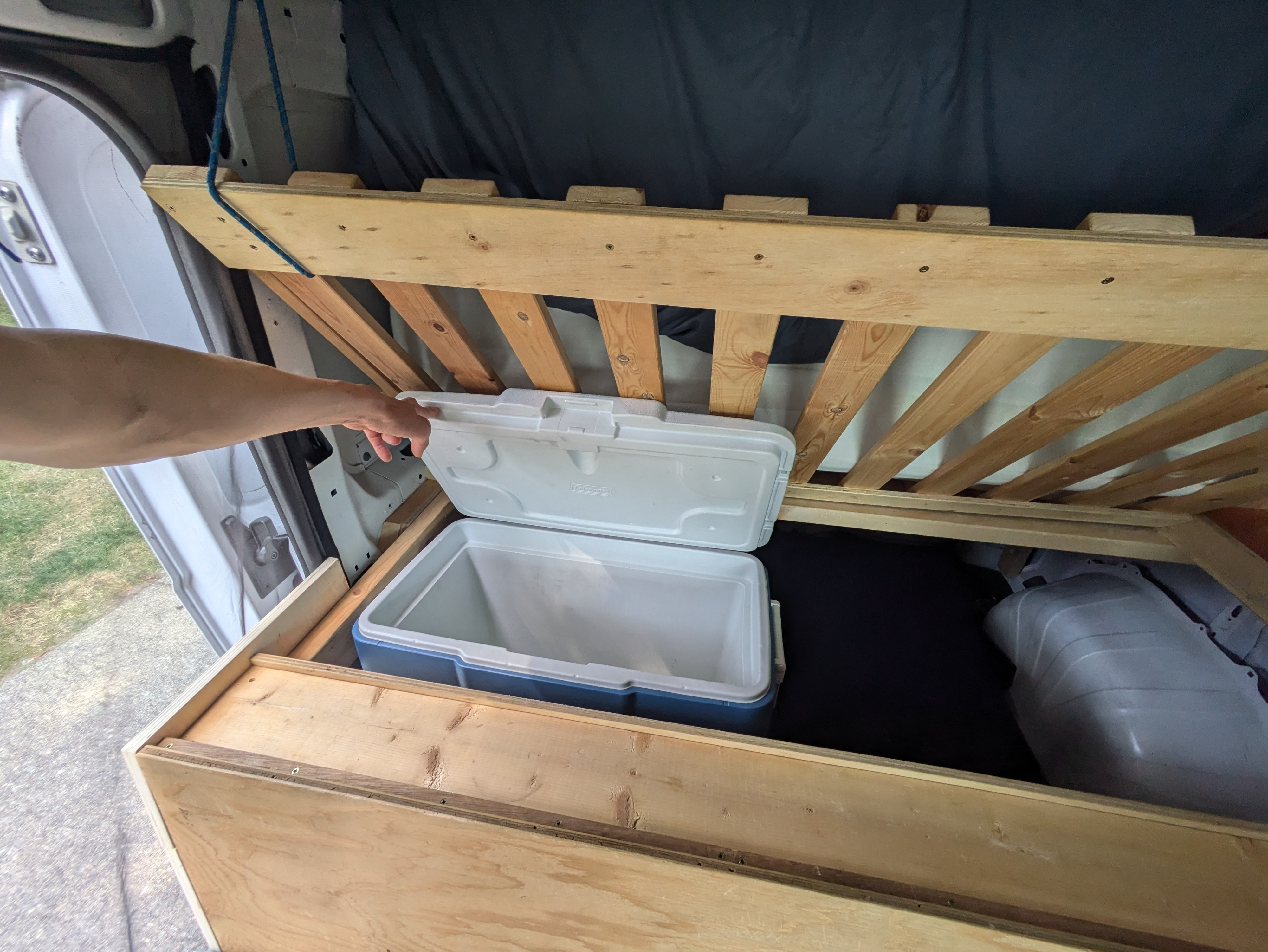 RV rental interior: Hand opening a cooler under a wooden slatted bed frame, showcasing efficient storage.. Dodge Ram Promaster 2017