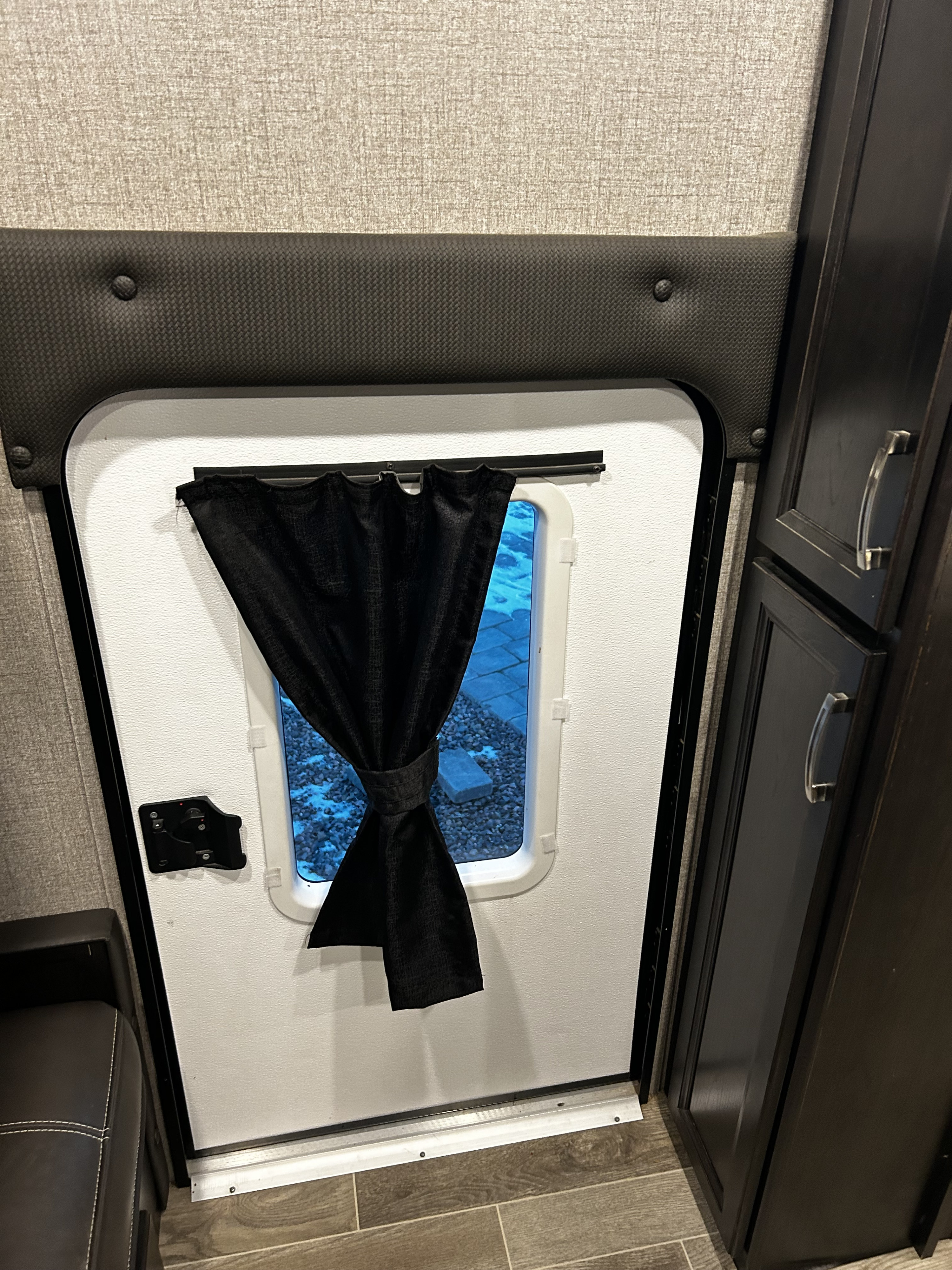 Interior of an RV rental: a white side door with a black, tied-back curtain. The window shows gravel and a blue paver outside.. Jayco Jay Feather 2021