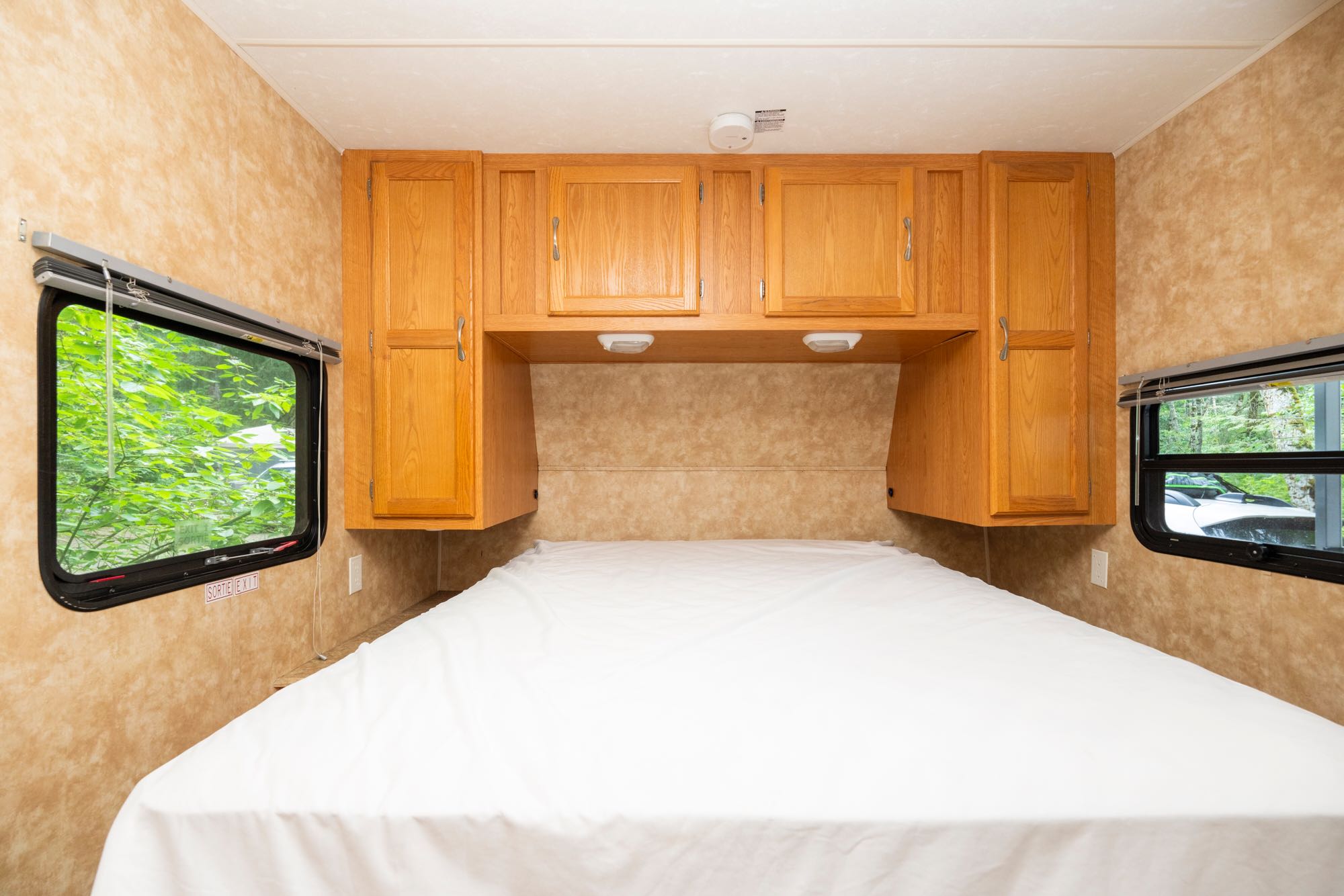Cozy RV rental bedroom with a comfortable bed, overhead cabinets, and windows showing green foliage outside.. Grand Haven Grand Haven Trailer 2008