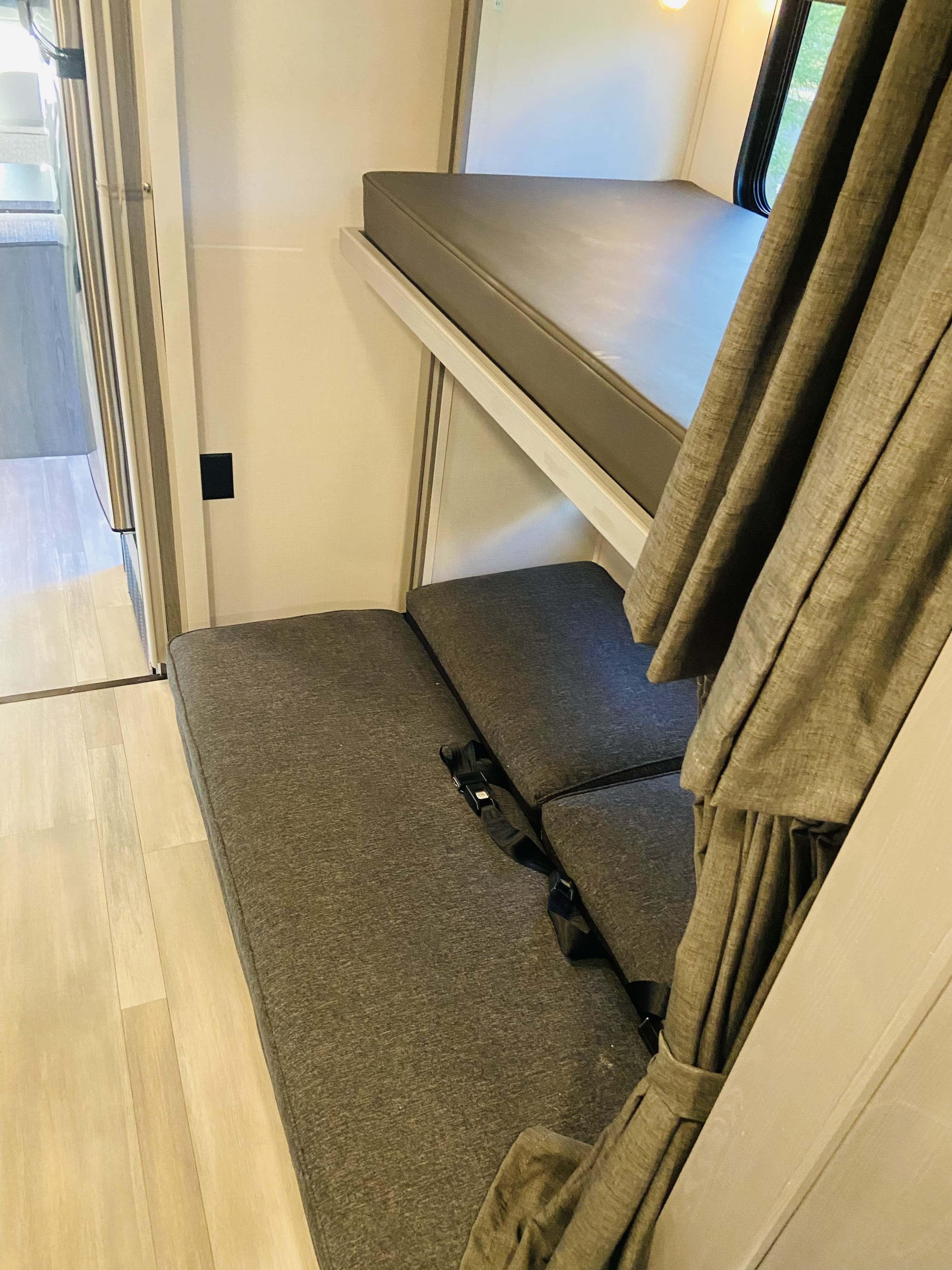 RV rental interior with an upper bunk bed, a lower seating area with safety belts, a curtain, and light wood flooring.. Forest River Solera 2023