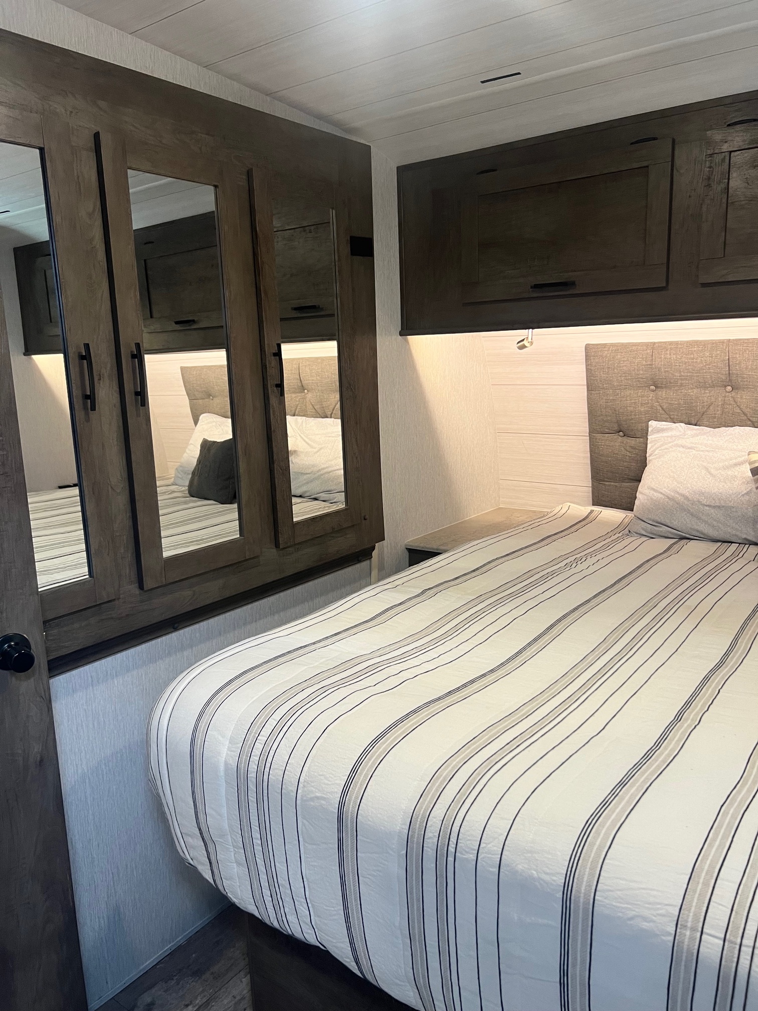 Cozy RV rental bedroom featuring a striped bed, mirrored wardrobe, and overhead cabinets for storage.. Forest River Sandstorm 2022