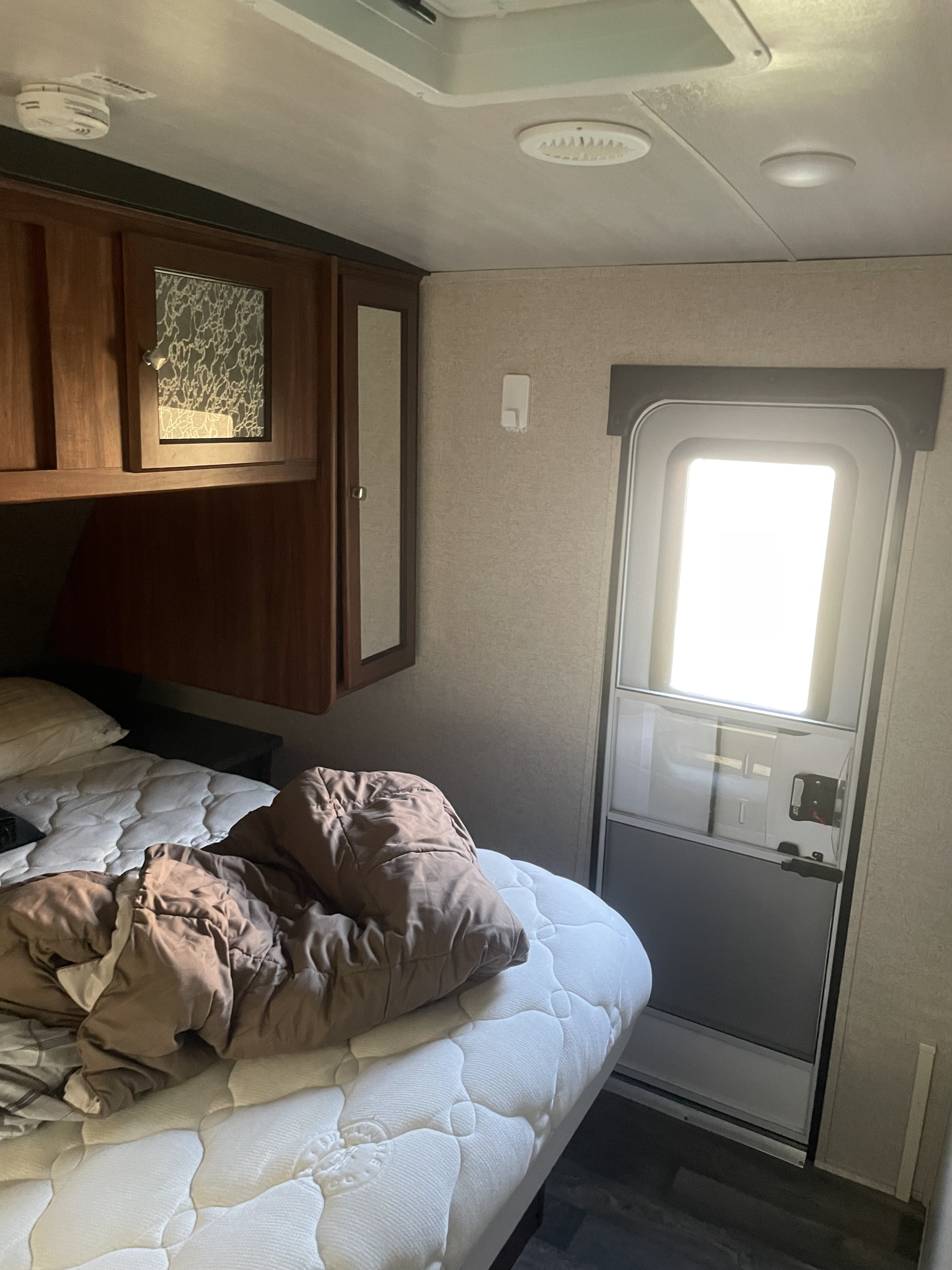 Interior of an RV rental bedroom showing a bed, wooden cabinets, and a bright main door. Ceiling has a smoke detector.. Heartland RVs Mallard 2017