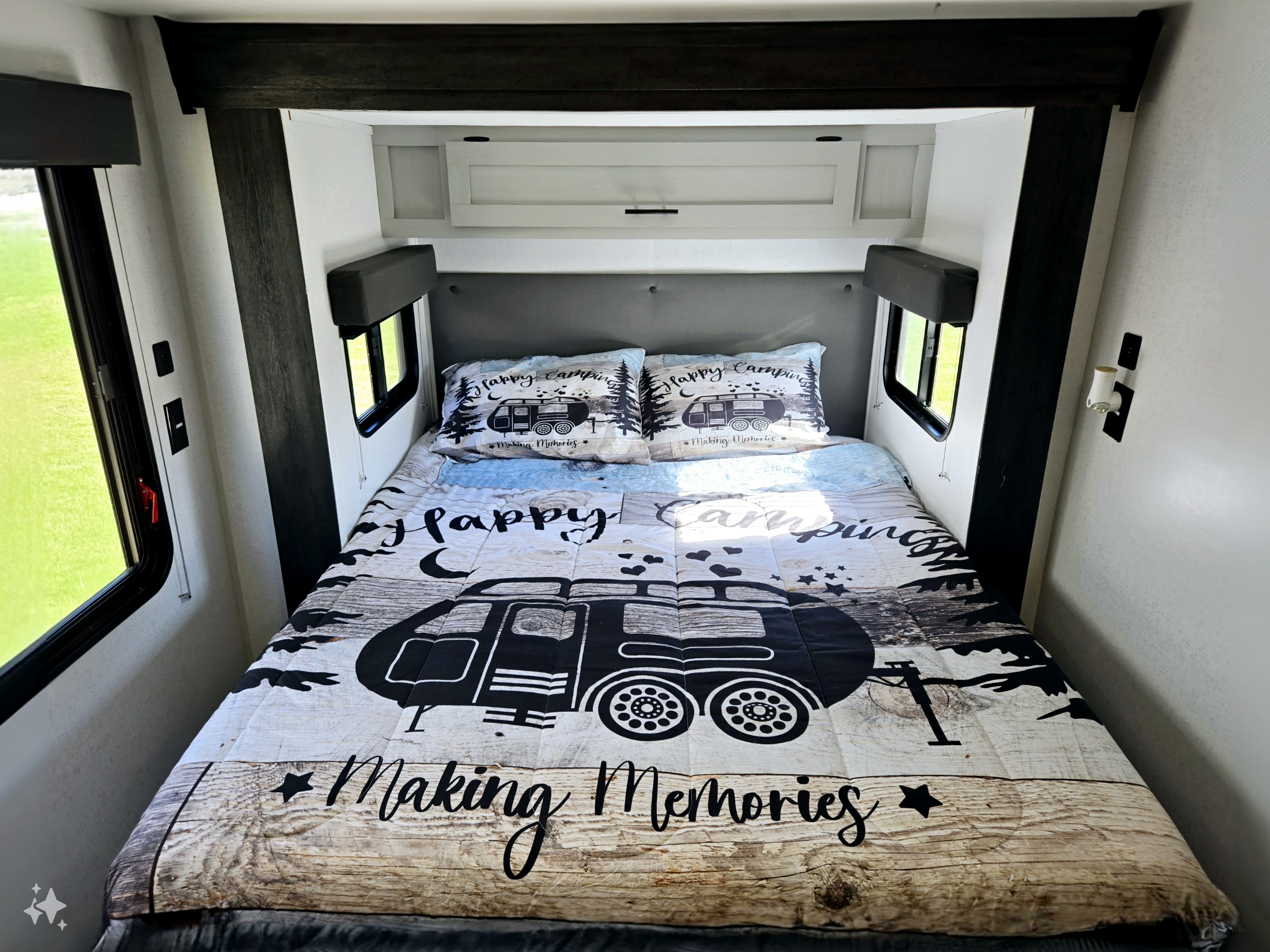 Cozy RV rental bedroom features a 'Happy Camping' themed bed, windows showing green outdoors, & overhead storage.. Forest River Wildwood FSX 2022