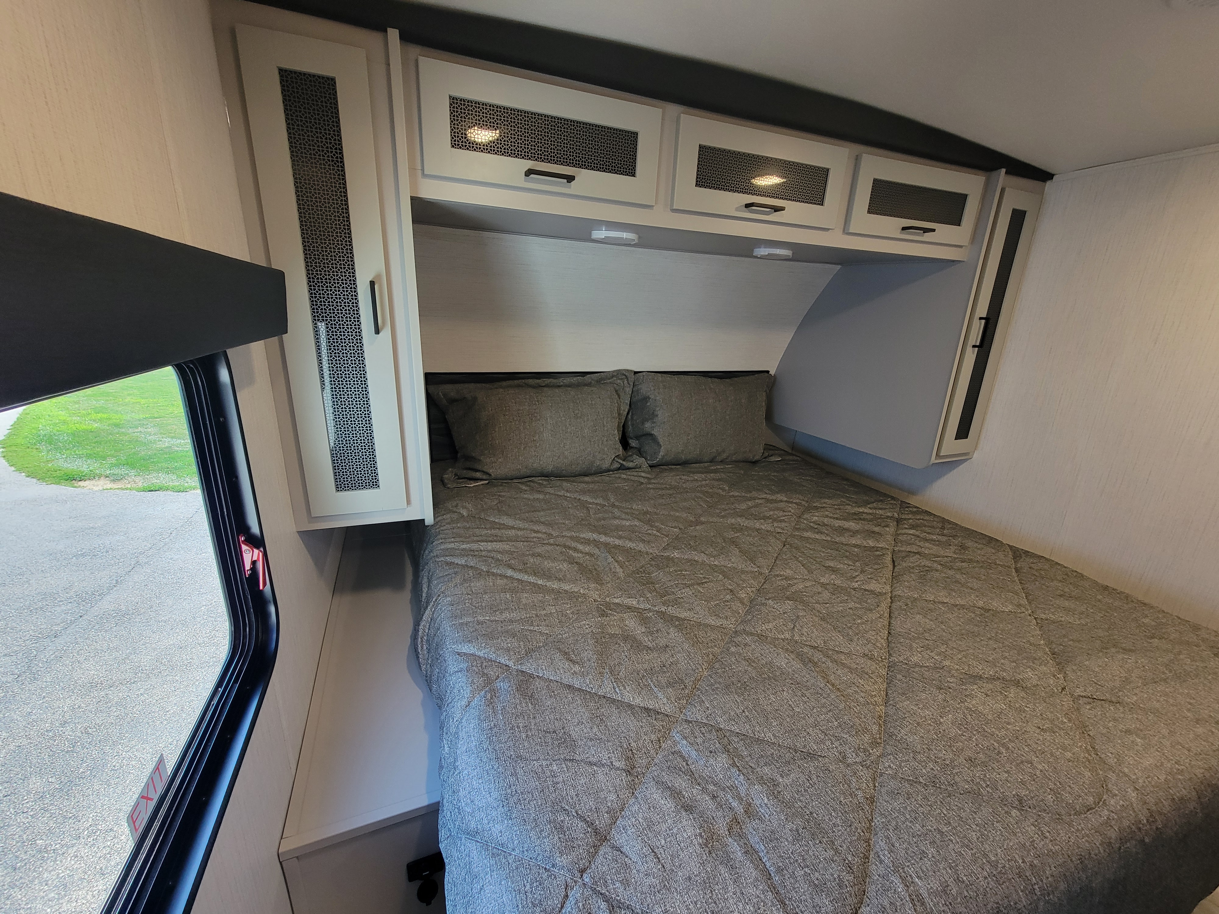 Cozy RV rental bedroom with a comfy grey bed, modern overhead cabinets, and a window offering outdoor views.. Heartland RVs North Trail 2023