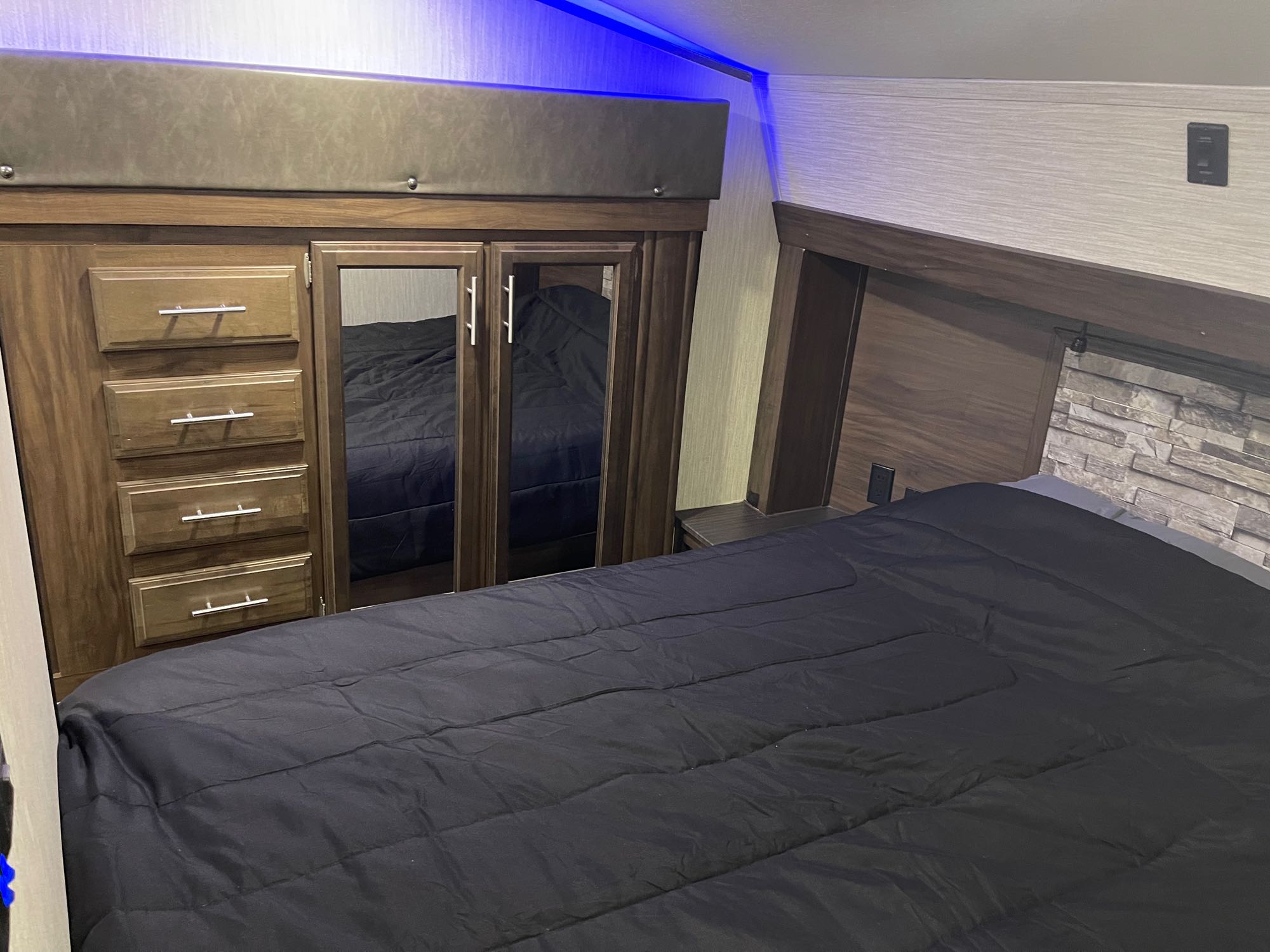 Cozy RV rental bedroom featuring a spacious black bed, built-in wardrobe, drawers, and modern stone accent wall.. Forest River Cherokee Arctic Wolf 2019
