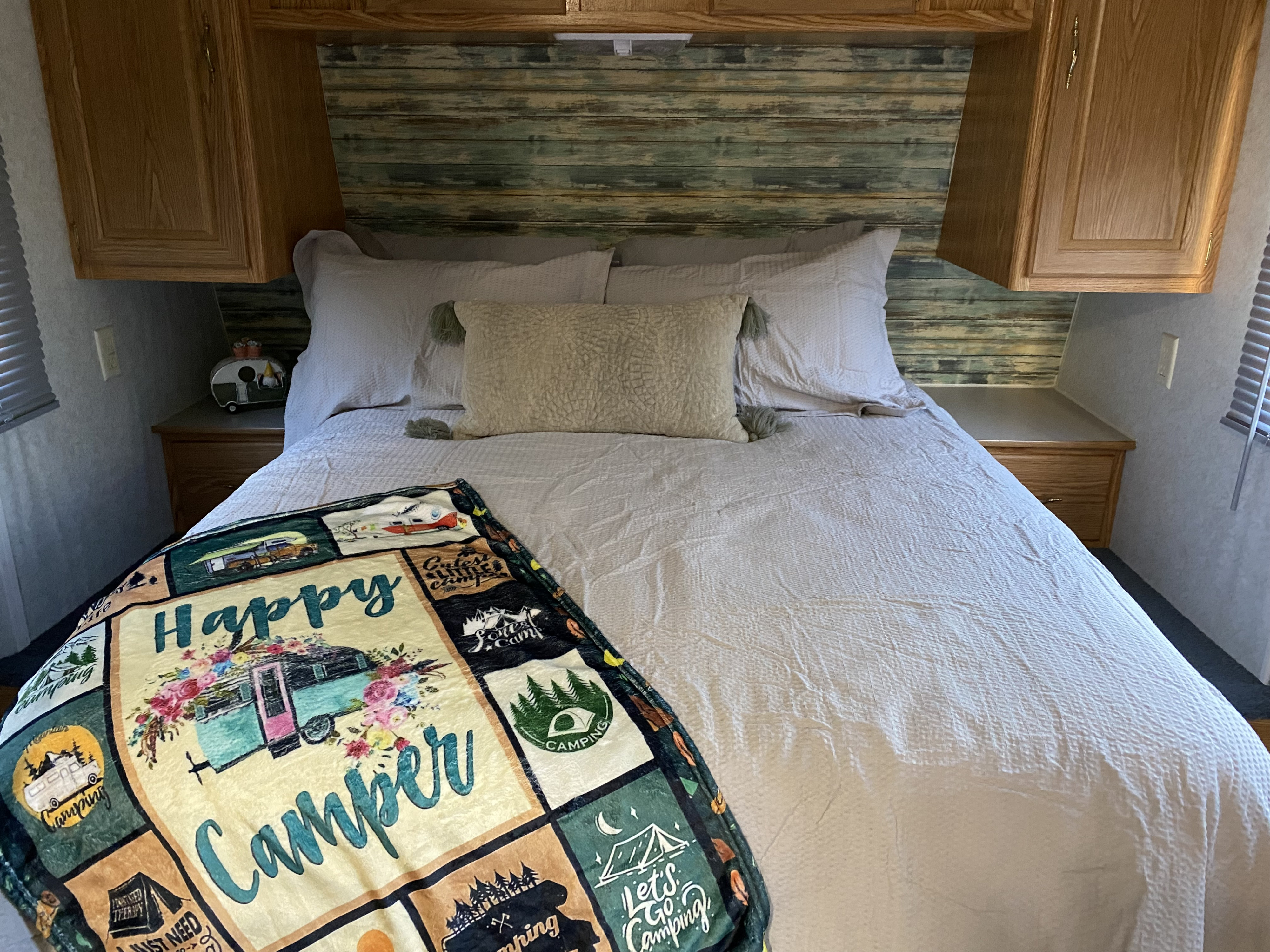 Cozy RV rental bedroom with a "Happy Camper" blanket, soft bedding, and rustic wood-paneled headboard.. Dutchmen Lite 2003