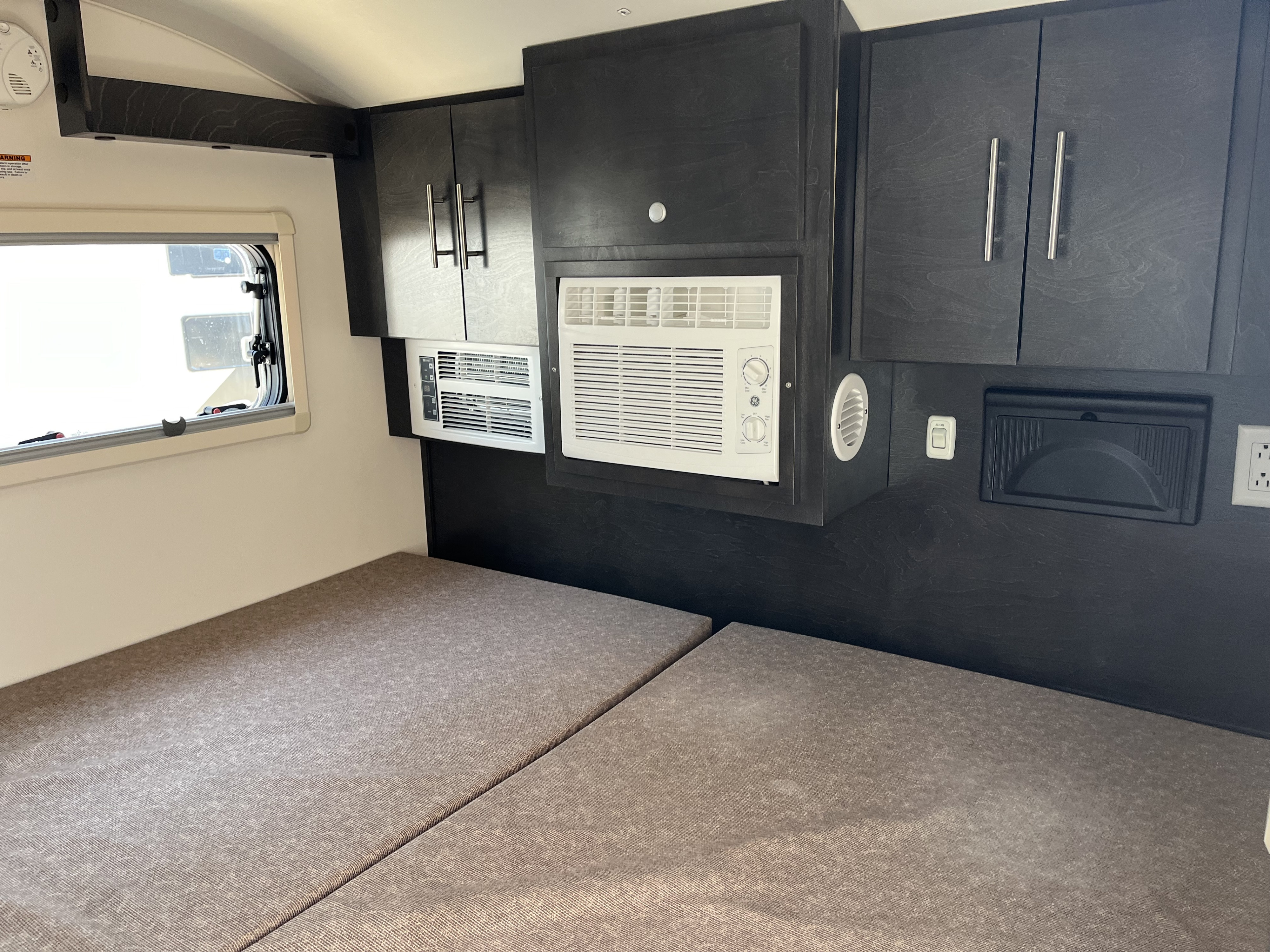 Inside an RV rental: a sleeping area with dark cabinets, a window, and two air conditioning units.. nuCamp T@G 2023