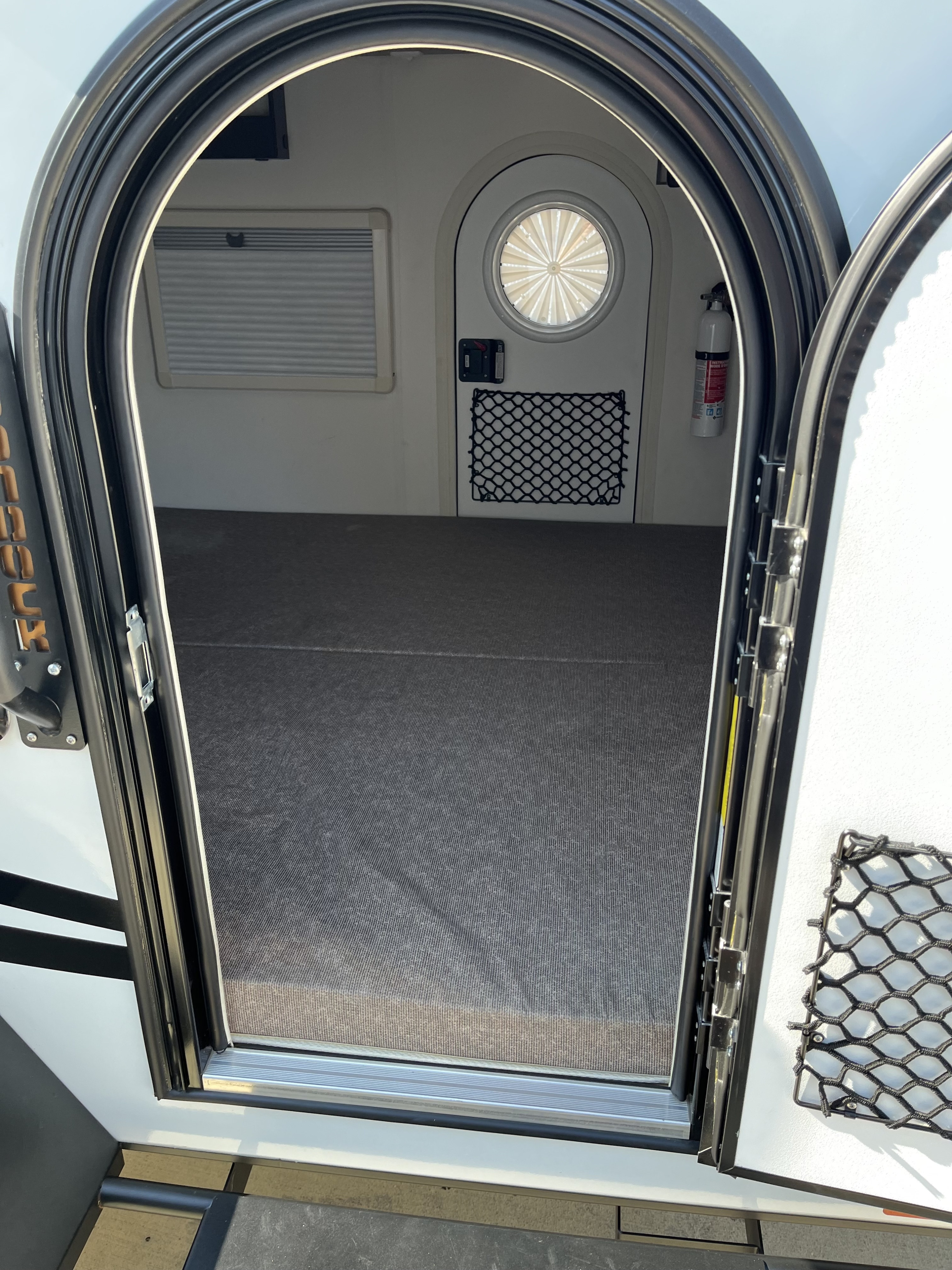 Inside a compact RV rental featuring an arched door, window blinds, fire extinguisher, and mesh storage net for travel.. nuCamp T@G 2023