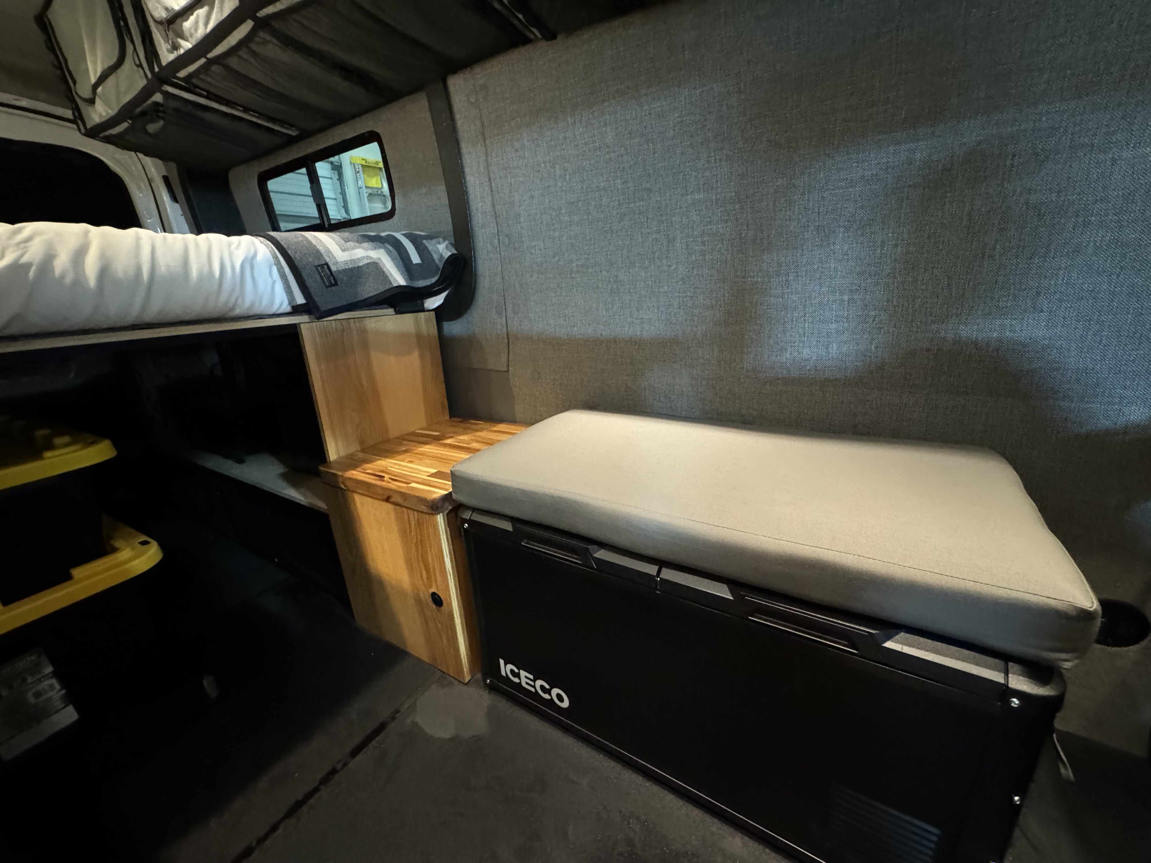 An RV rental's versatile interior features a bed, ICECO portable fridge with seating, and wood storage.. Ford Transit Custom 2023