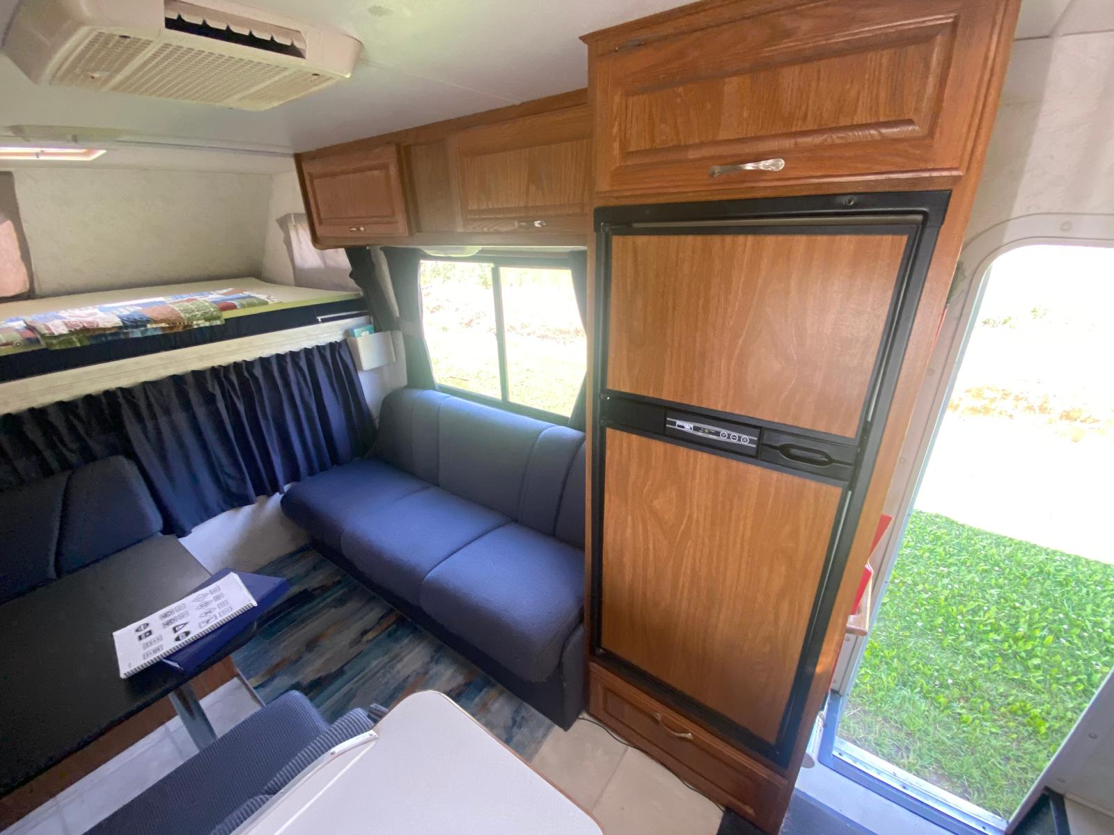 Comfortable RV rental interior showing a bunk bed, blue sofas, dinette, and wood-paneled fridge. Ideal for travel.. Thor Majestic 19G 2004