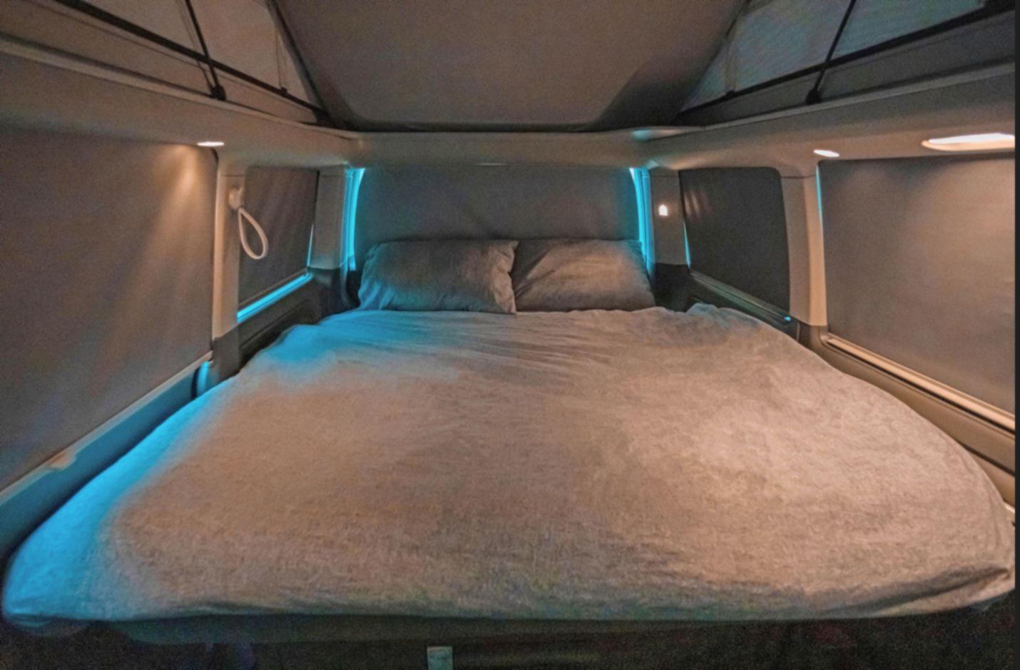 Experience a cozy RV rental bedroom. This inviting space features a comfortable bed with ambient blue LED lighting and neutral tones.. Volkswagen California Beach Camper 2024