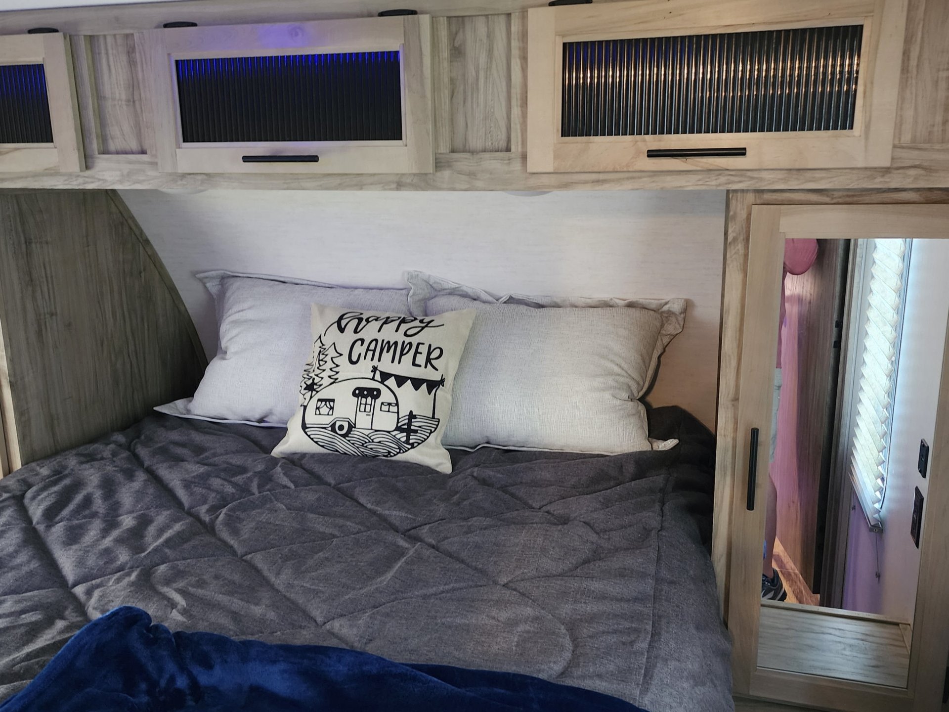 This RV rental offers a cozy bedroom with a grey comforter, "Happy Camper" pillow, and lighted overhead cabinets.. Forest River Cherokee 2022