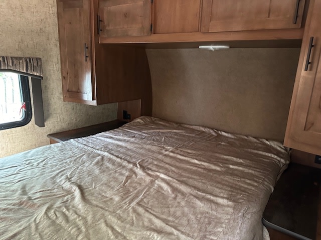Inside a comfortable RV rental, a neatly made bedroom features an inviting bed, ample overhead cabinets, and a scenic window.. Outdoors RV Timber Ridge 2020