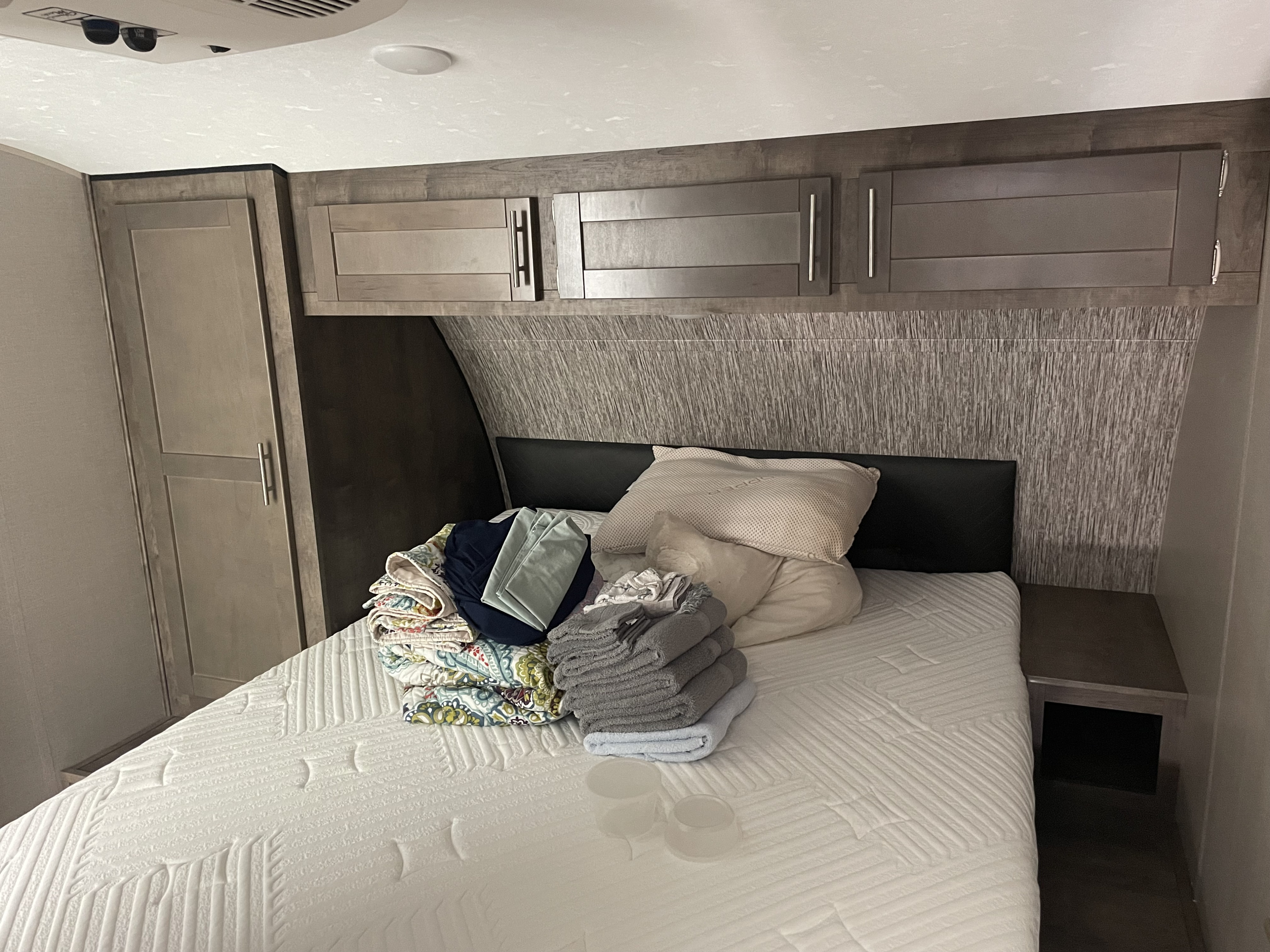 A tidy rv rental bedroom featuring a bed with stacked bedding and towels, built-in overhead cabinets, and a wardrobe.. Connect Connect Trailer 2020