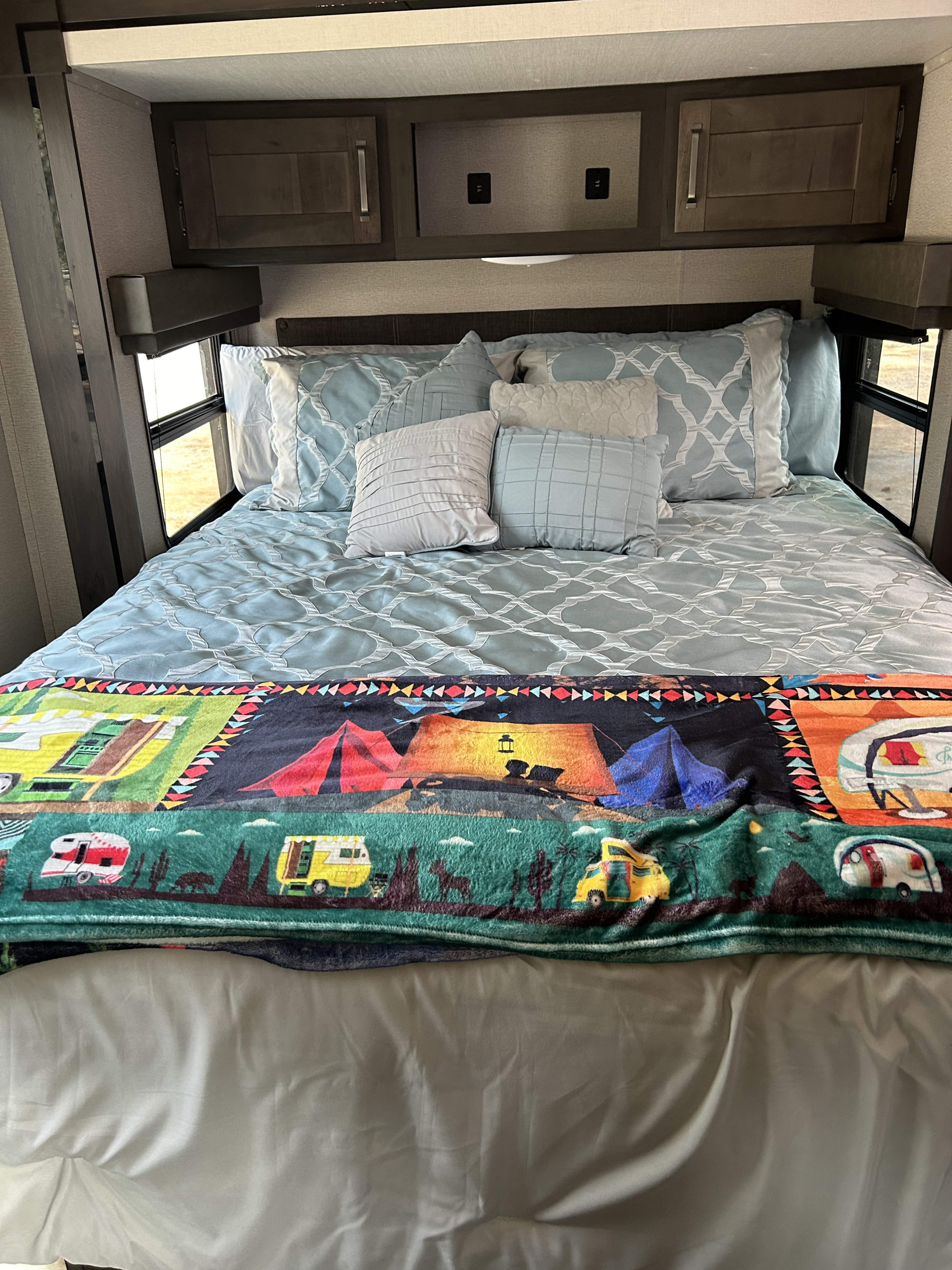 Cozy RV rental bedroom with patterned bedding, a colorful camping blanket, overhead storage, and side windows.. Grand Design Transcend Xplor 2023