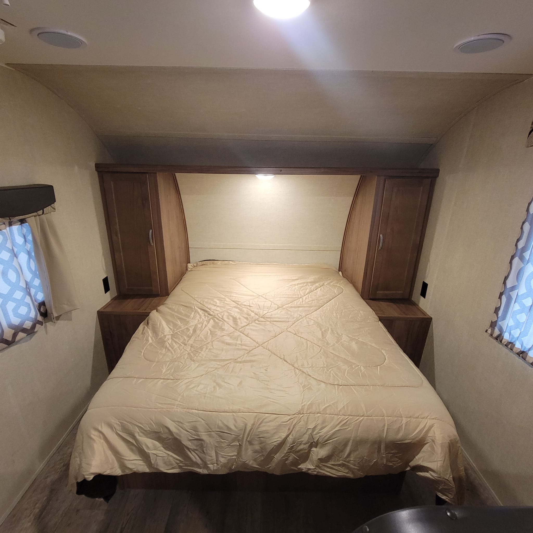 RV rental bedroom: a spacious bed with light comforter, flanked by cabinets, under bright ceiling lights. Cozy travel space.. Gulf Stream Ameri-Lite 2018