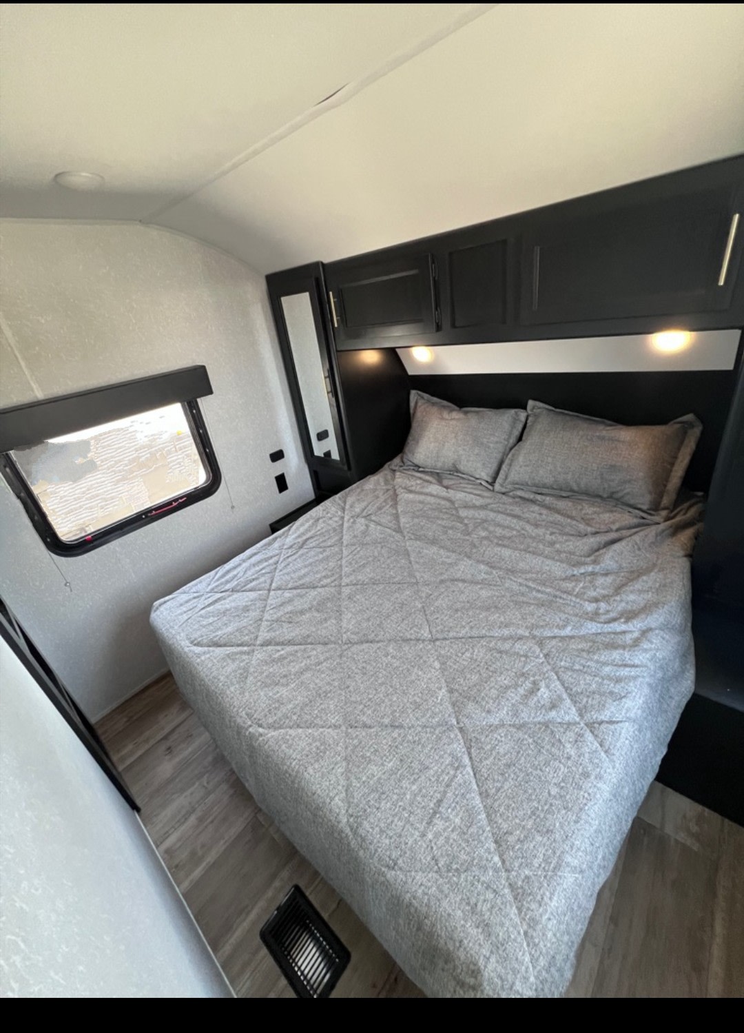Cozy RV rental bedroom featuring a grey bed with pillows, dark overhead cabinets, a side window, and wood-look flooring.. Forest River Cherokee 2022