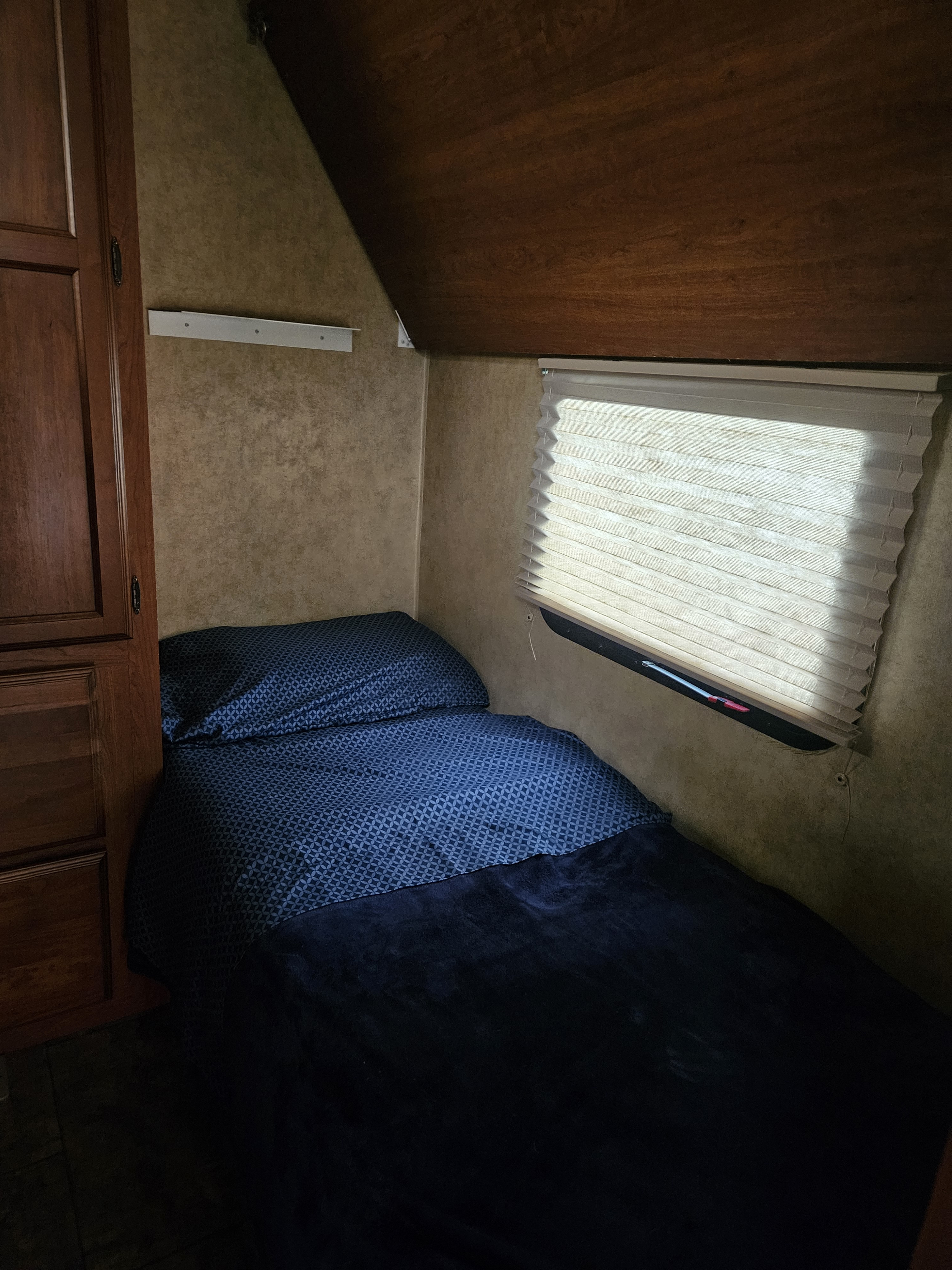 A compact RV rental bedroom with a neatly made bed featuring blue patterned sheets and a plush blanket, alongside a window with a pleated shade.. Dutchmen Aerolite 2013
