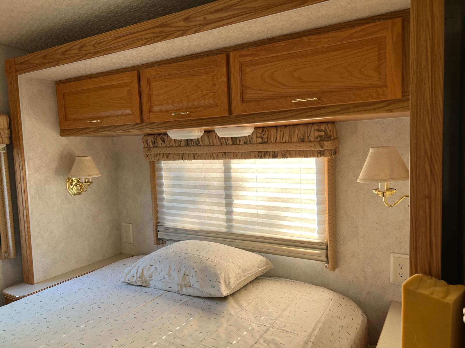 RV rental bedroom: wooden cabinets, bed with patterned sheets, window blinds, and wall sconces.. Itasca Spirit 2003