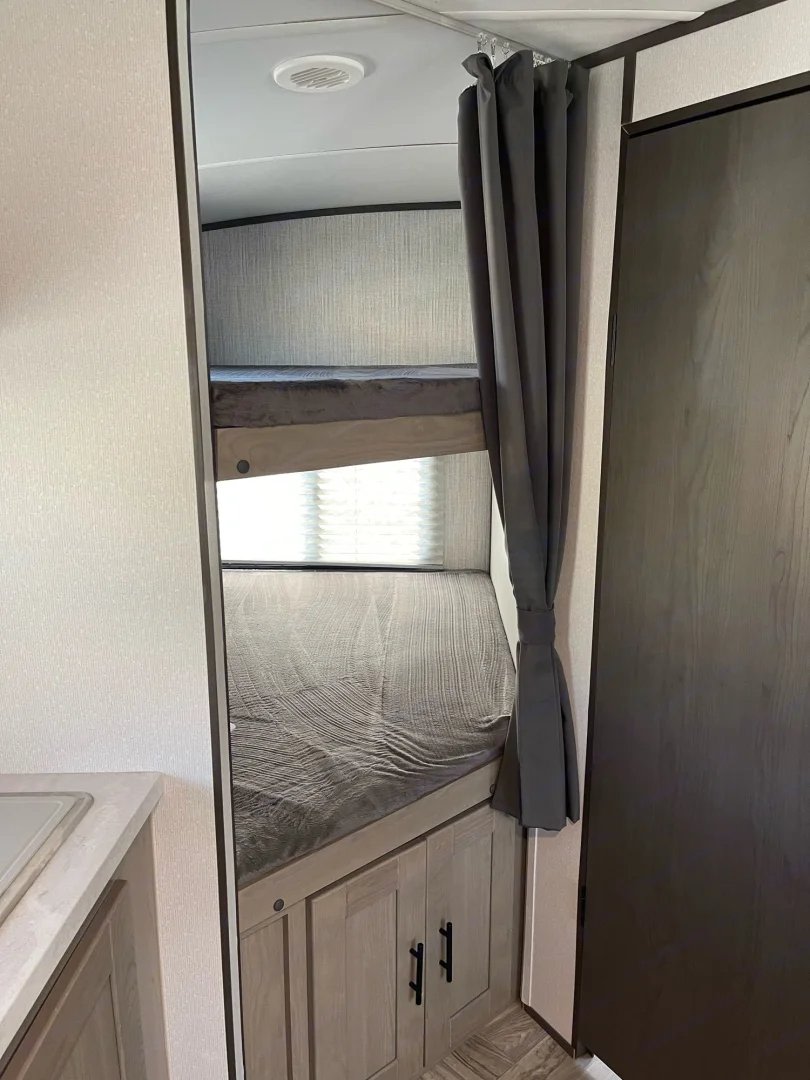 RV rental interior: two gray bunk beds, a light wood kitchen counter, and lower cabinets. A dark curtain is tied back.. Cruiser RV Shadow Cruiser 2022