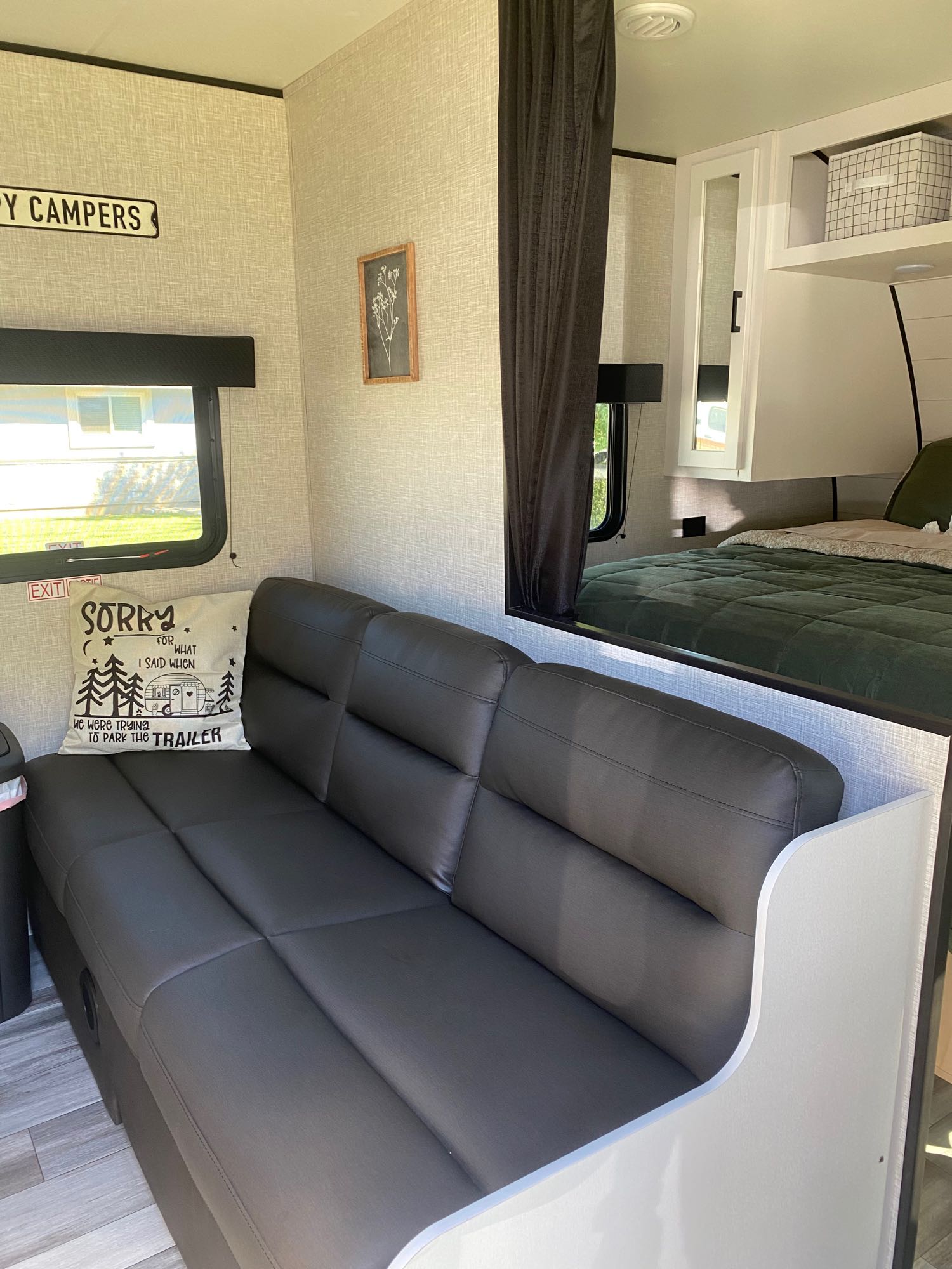 Inside a modern RV rental: gray sofa, raised bed with green comforter, and "Happy Campers" decor.. Jayco Jay Flight SLX 2024