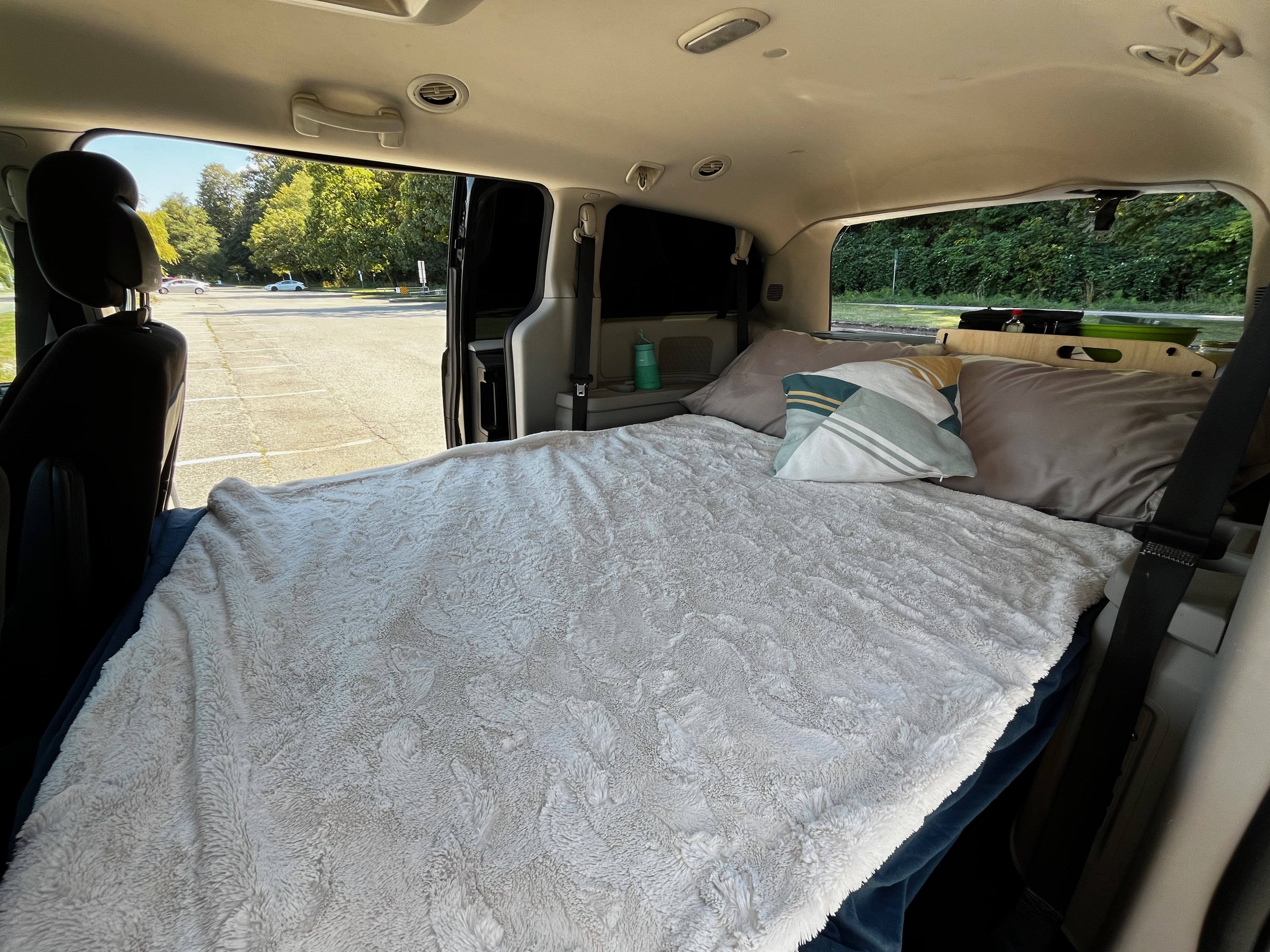 Inside a cozy minivan, converted for an RV rental. Features a comfy bed, pillows, and an outdoor view of a sunny parking lot.. Dodge Grand Caravan 2014