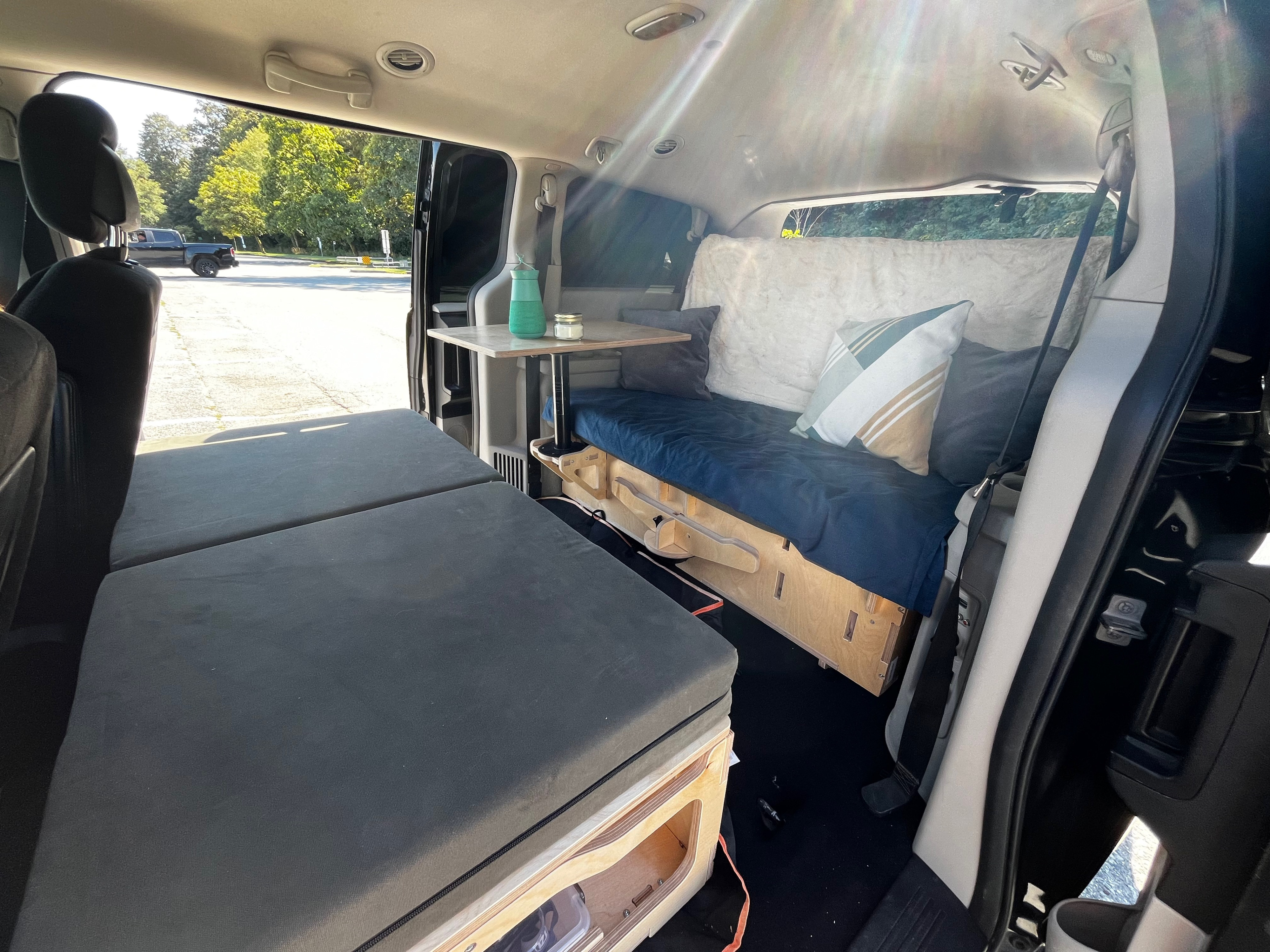 A versatile RV rental: this minivan conversion features a long gray bed, a blue bench seat with pillows, and a small table.. Dodge Grand Caravan 2014