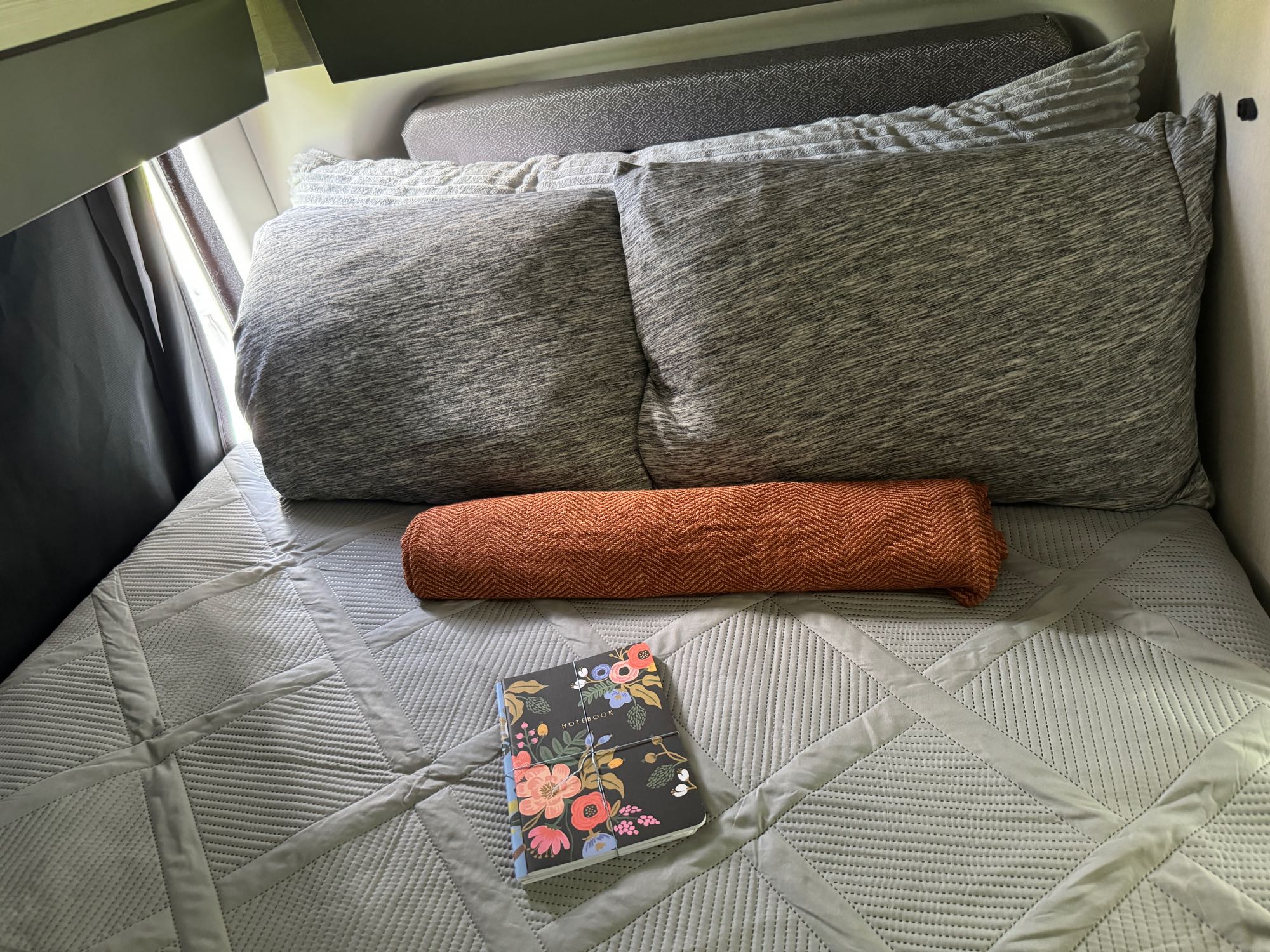 Inviting RV rental bed with soft gray pillows, a chevron-patterned orange blanket, and a floral notebook.. Thor Sanctuary 2024