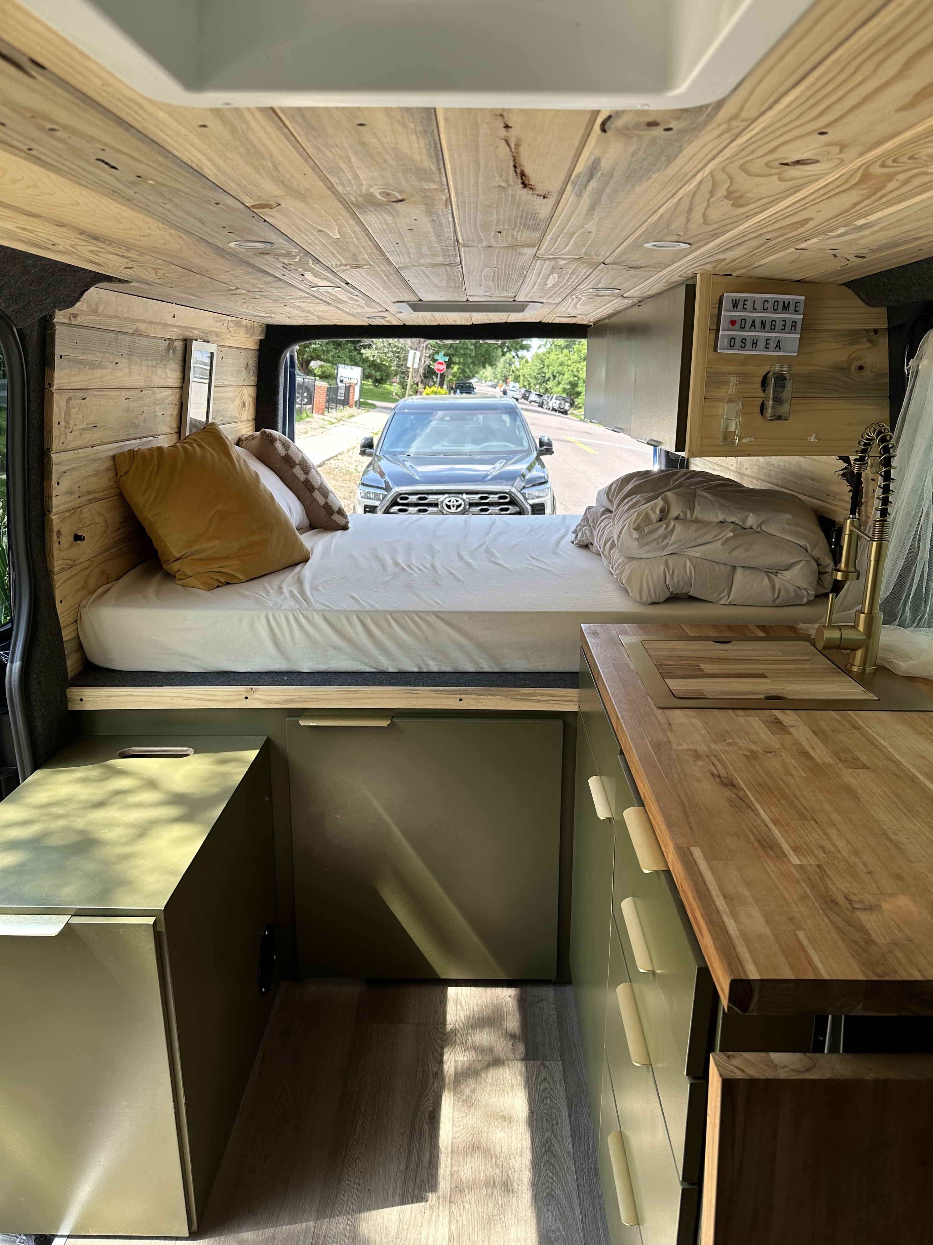 Cozy custom RV rental interior with wooden bed area, olive green storage, compact kitchen, and street view.. Mercedes-Benz Sprinter 2016