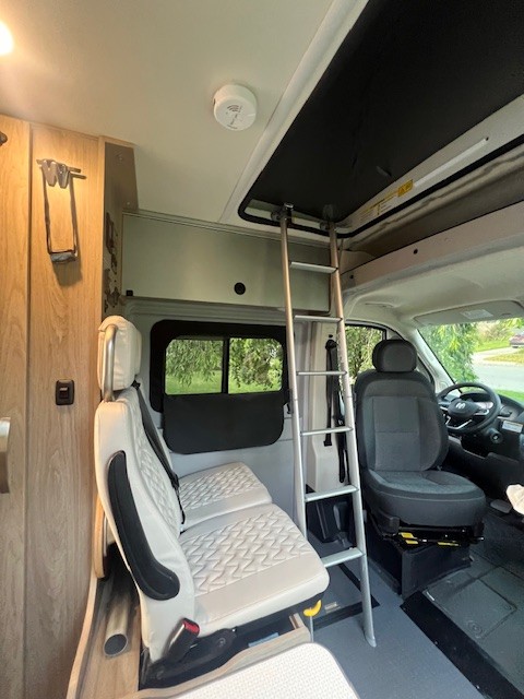Inside an RV rental: a modern camper van interior with front seats, a ladder to the overhead bunk, and wood accents.. Winnebago Solis 2023