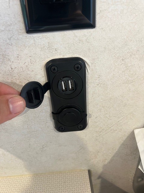 Hand holds open the cover of a dual USB charging port, a useful amenity for an RV rental.. Winnebago Solis 2023